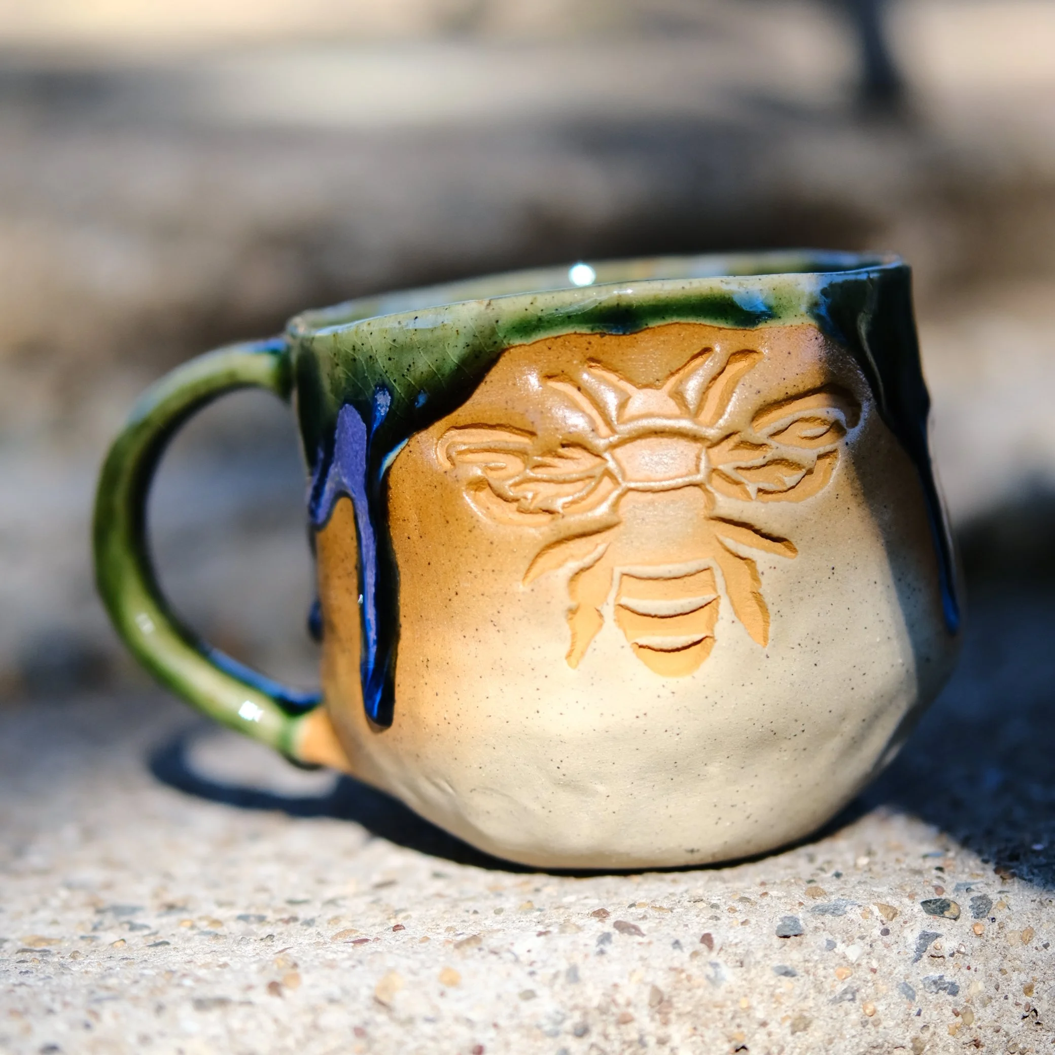 Handmade sand-colored ceramic mug with a mustard yellow bee motif and a drip-style green and purple glaze, displayed on a rustic background.