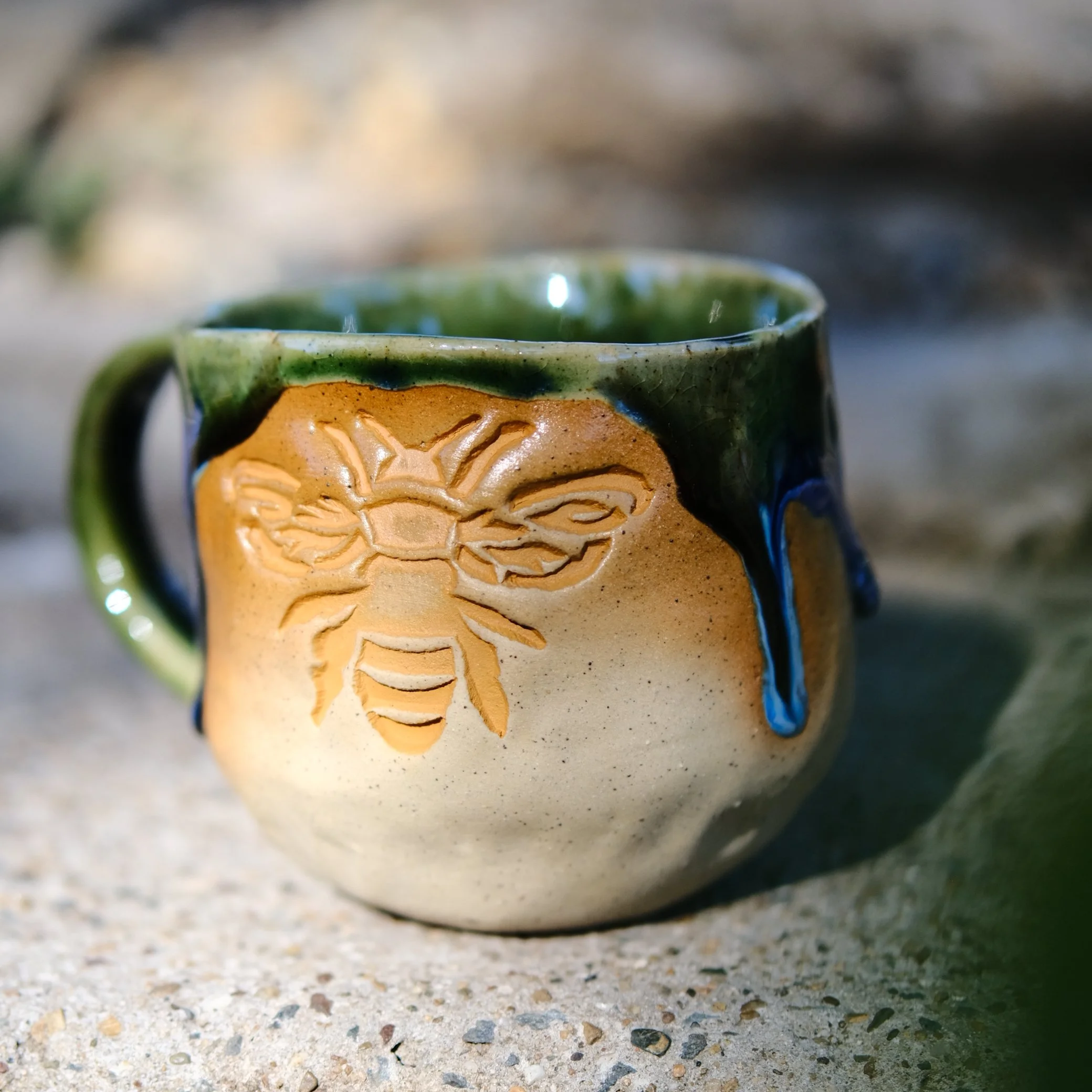 Handmade sand-colored ceramic mug with a mustard yellow bee motif and a drip-style green and purple glaze, displayed on a rustic background.