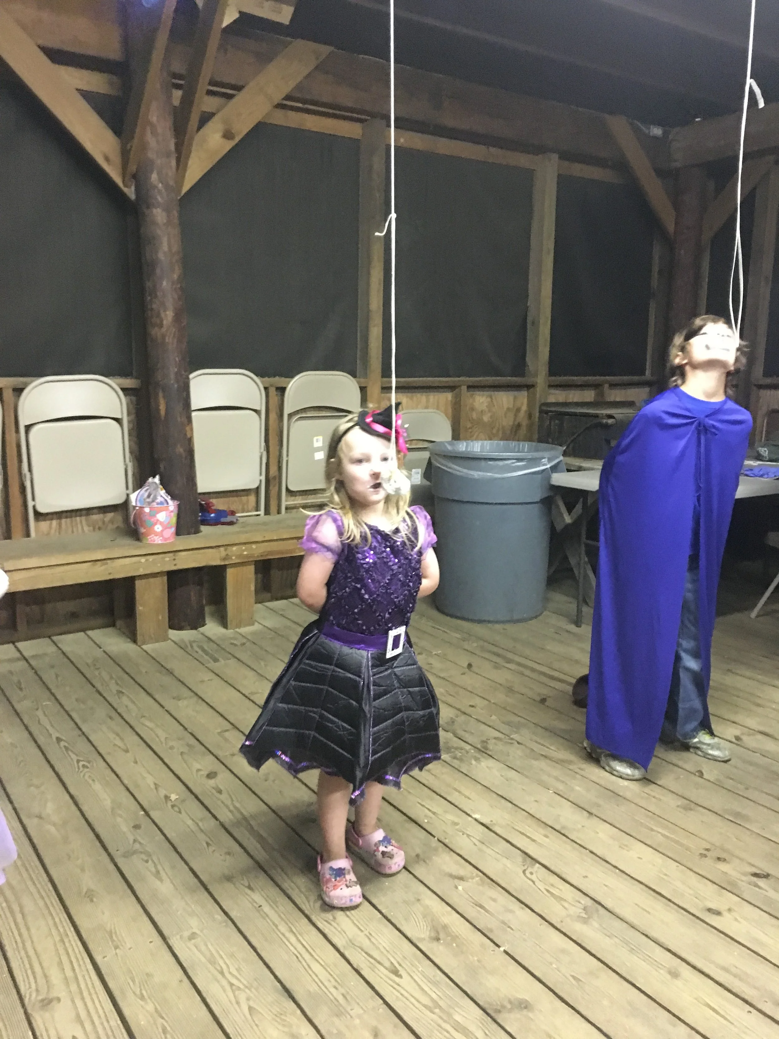 Two children in costumes are standing in a wooden indoor space, with one girl dressed as a purple princess and the other as a blue wizard.