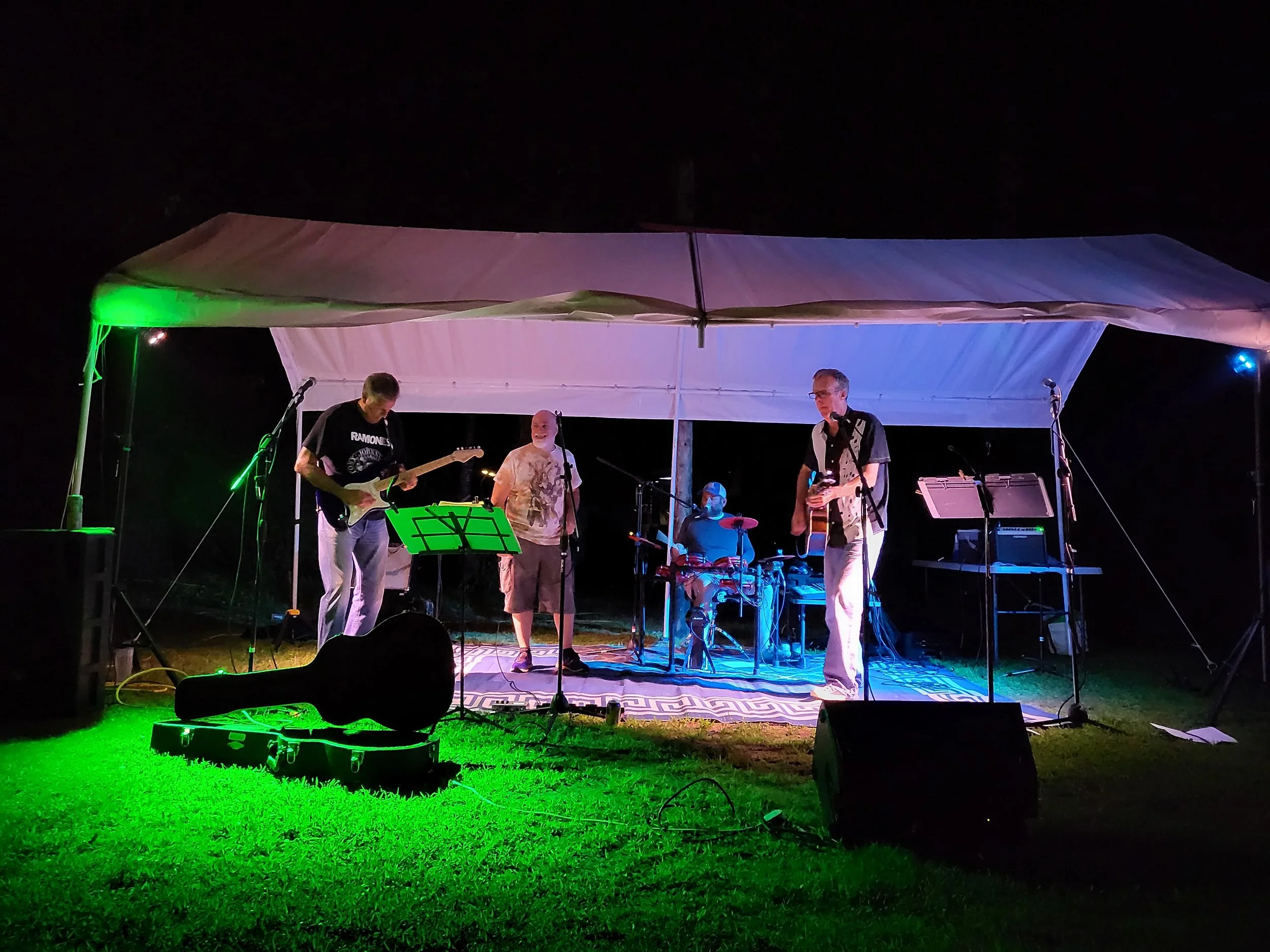 A band performing live on a small outdoor stage at night, illuminated by colorful stage lights, with four musicians playing guitars, drums, and singing.