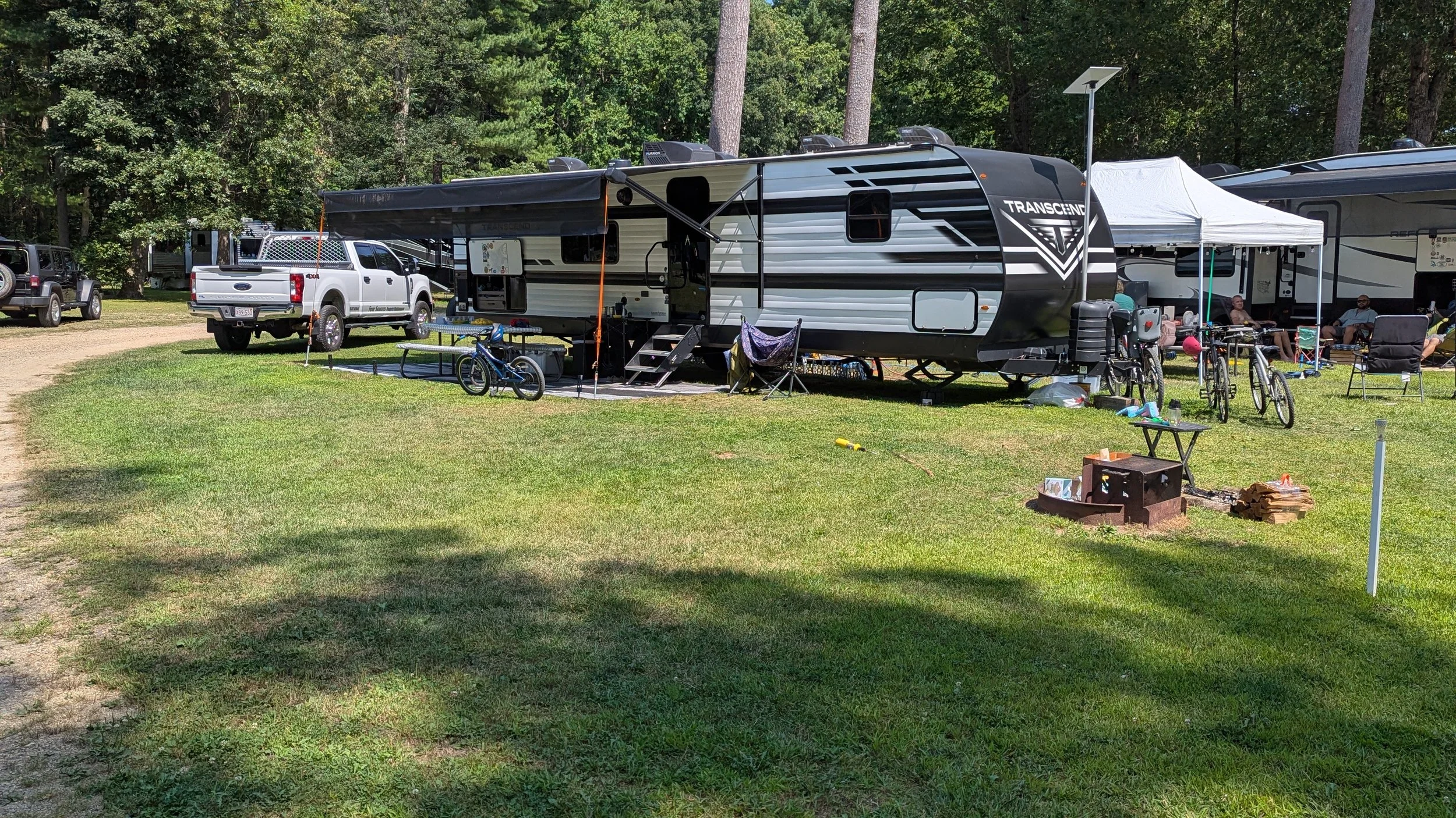 A campsite with a white and black RV, a white truck, bicycles, and outdoor furniture set on a grassy area surrounded by trees.
