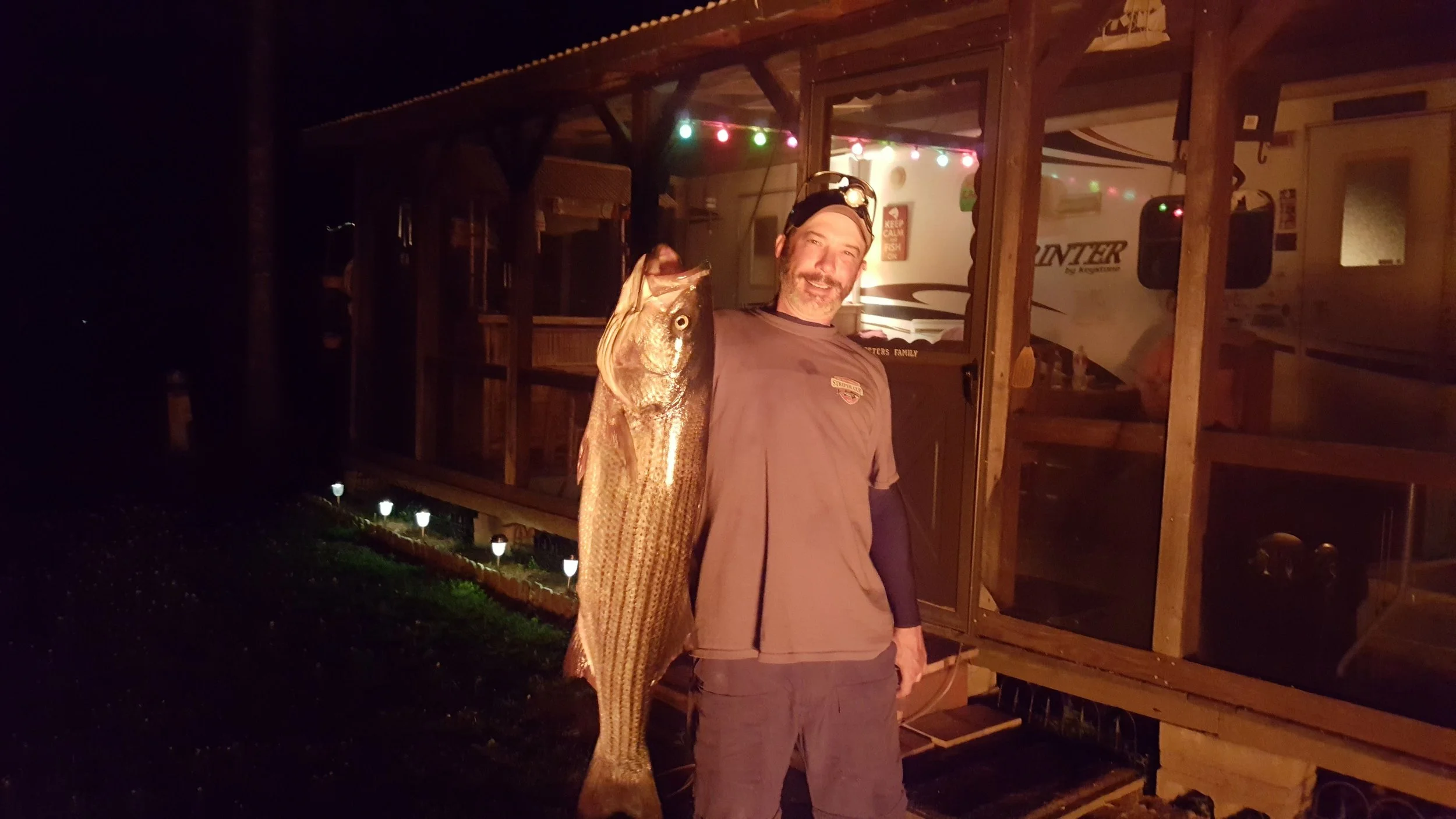 Man holding large fish in outdoor nighttime setting with a wooden porch and string lights in the background.