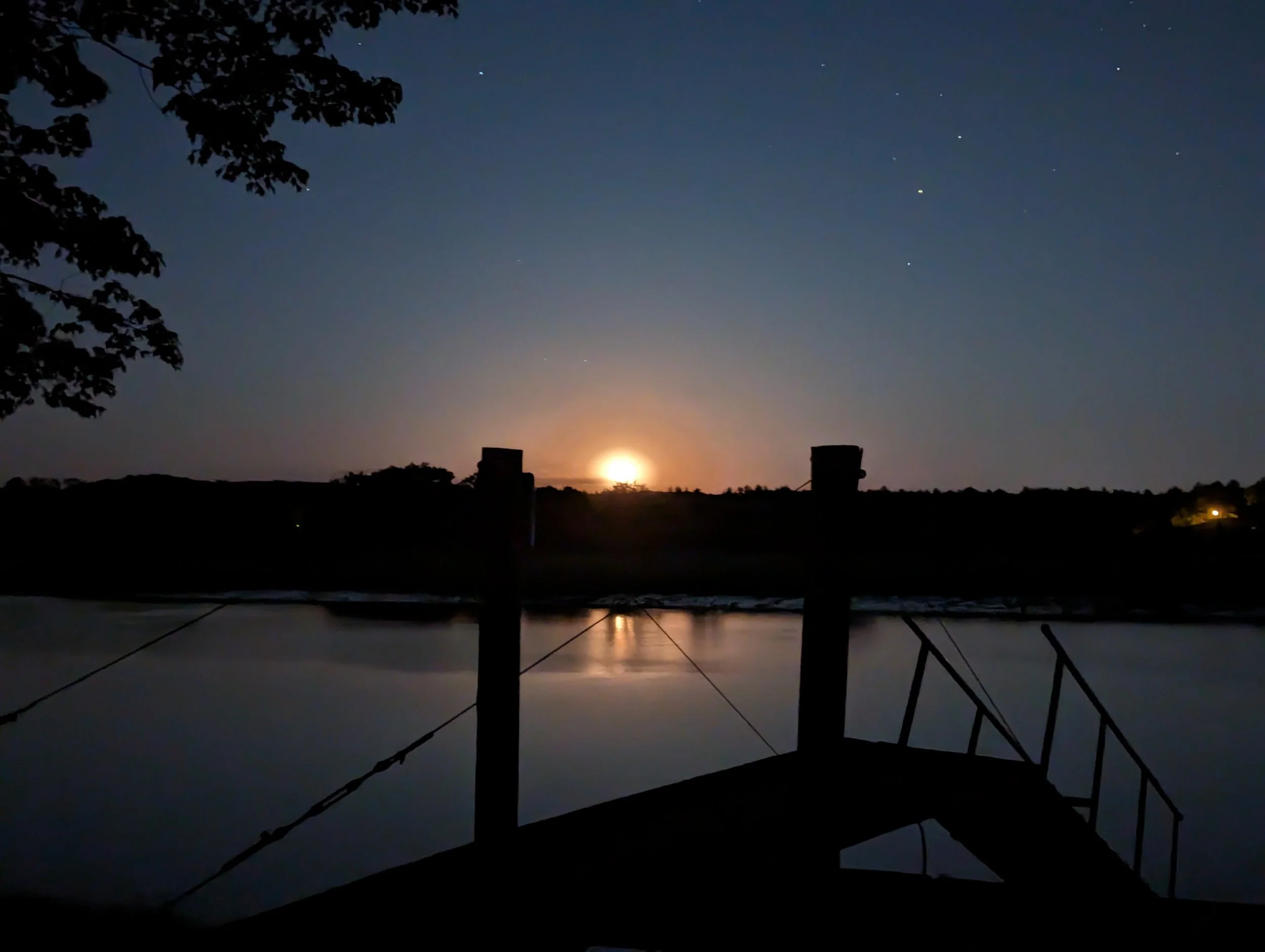Nighttime scene with a boat dock, calm water, trees, and a bright moon on the horizon with a few stars visible in the sky.