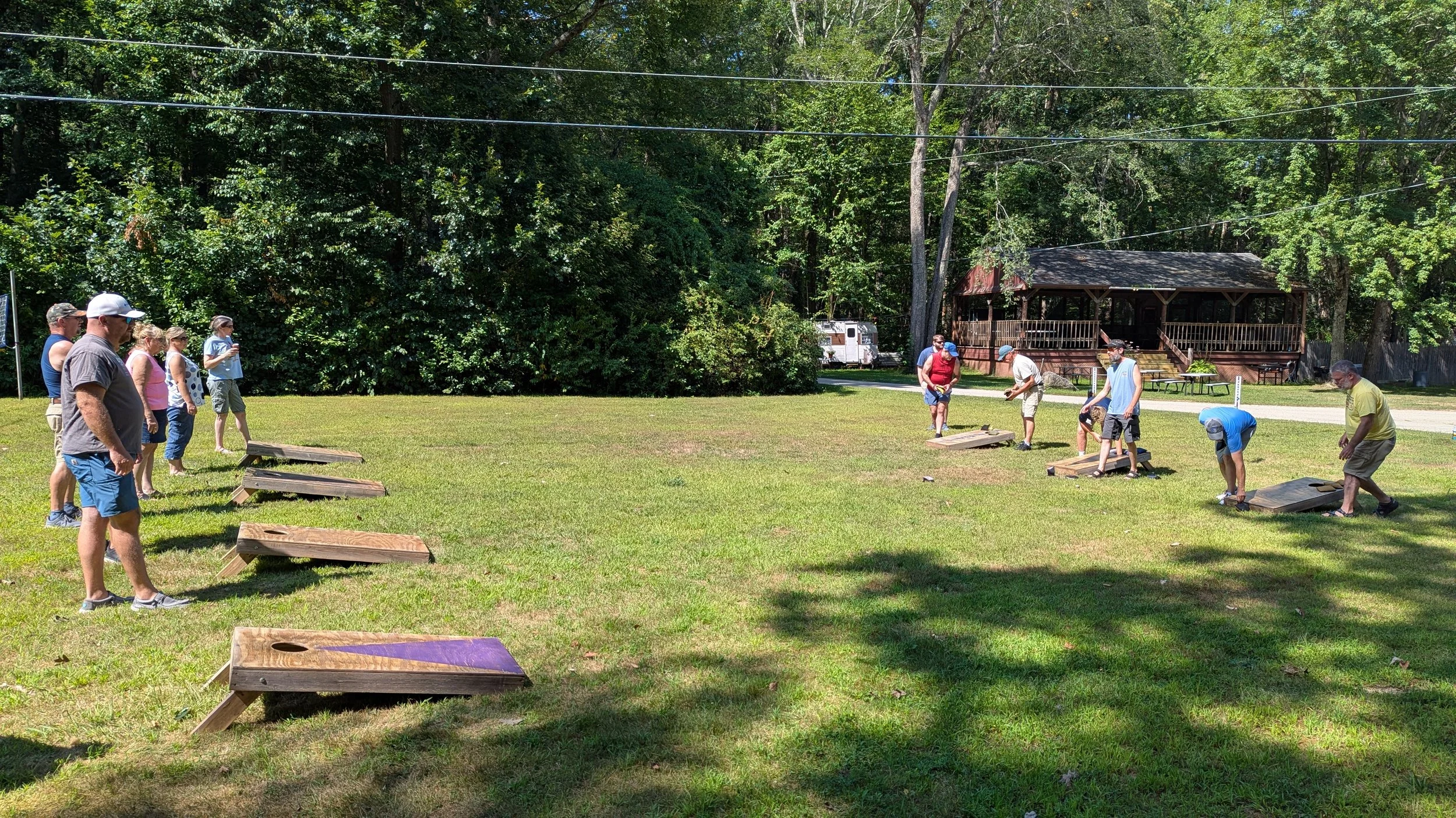 People playing cornhole on a grassy outdoor area surrounded by trees and a wooden cabin.