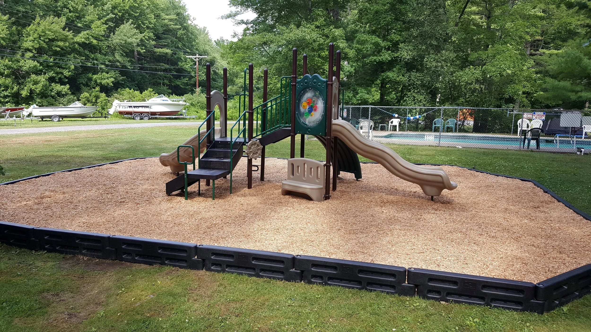 Children's playground with slide, climbing structure, and sandbox, surrounded by a black border and gravel surface, with boats on trailers and lush green trees in the background.