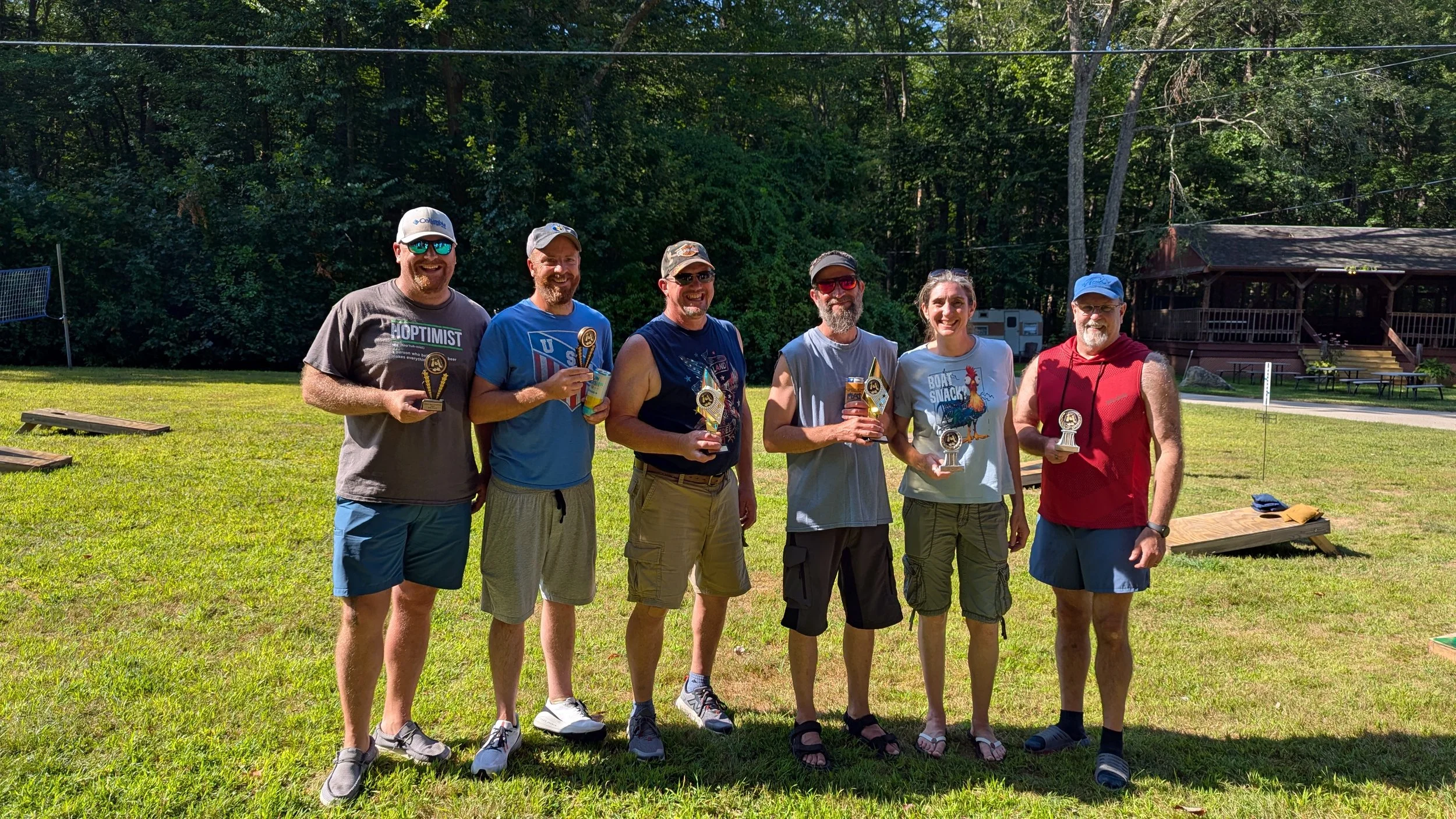Group of seven men standing on a grassy field holding awards, smiling, with trees and a wooden pavilion in the background.