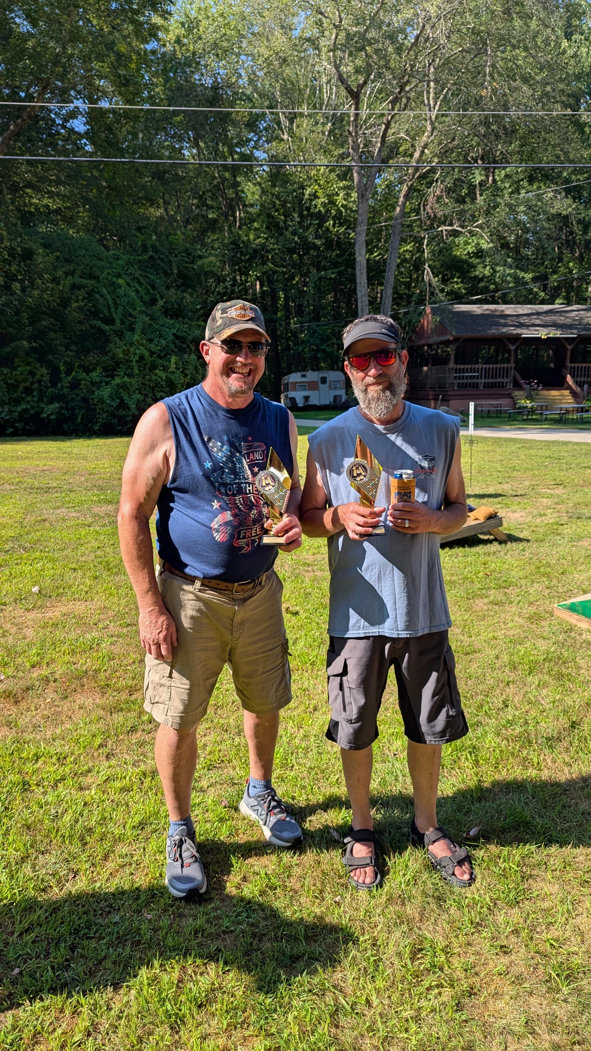 Two men standing outdoors on grass, smiling, holding trophies and a beverage can. They are dressed casually in sleeveless shirts, shorts, and sunglasses, with a background of trees, a wooden building, and a trailer.