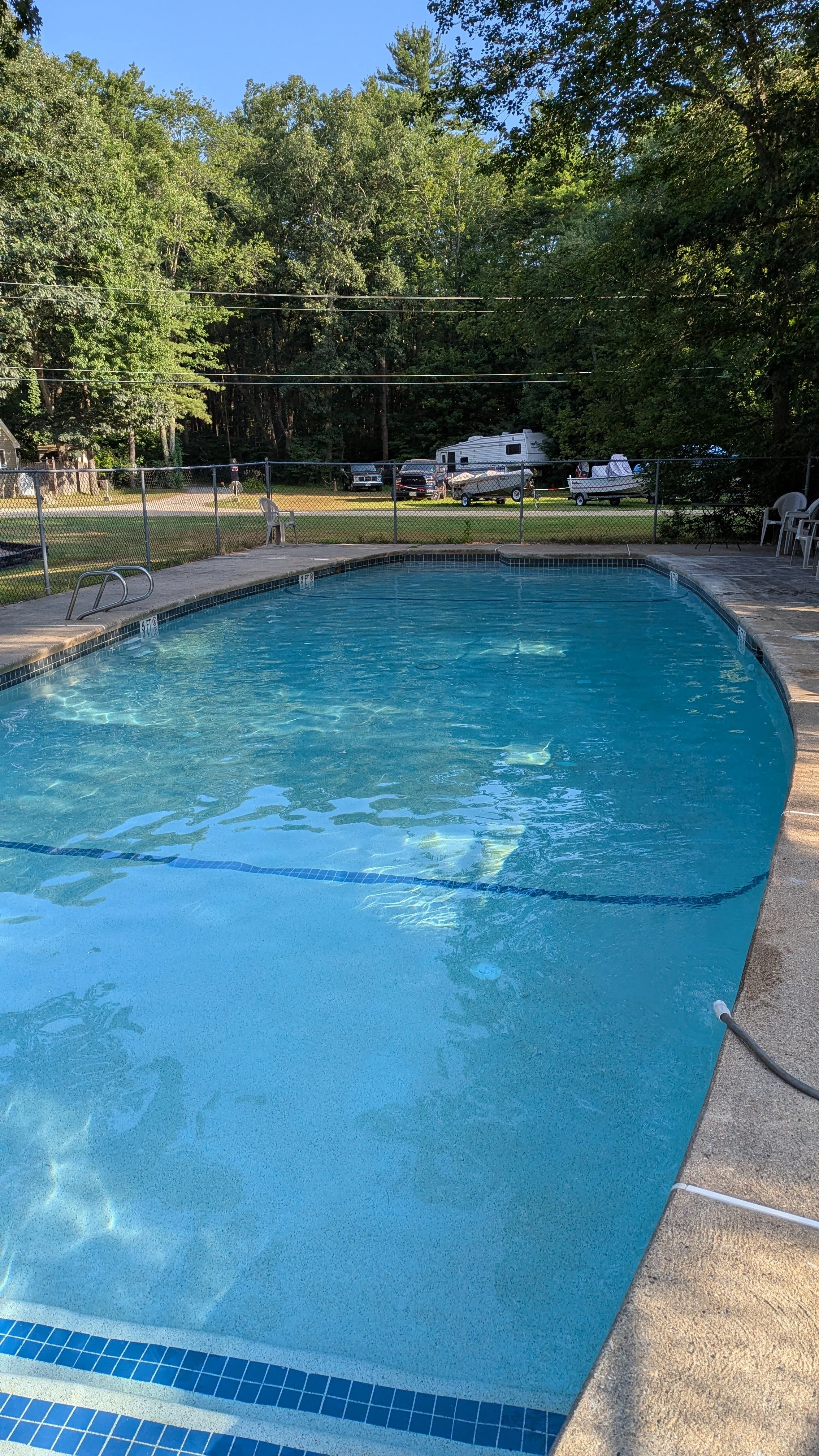 An outdoor swimming pool with blue water, surrounded by a concrete deck and a chain-link fence. In the background, there are trees, a grassy area, parked cars, and a trailer.