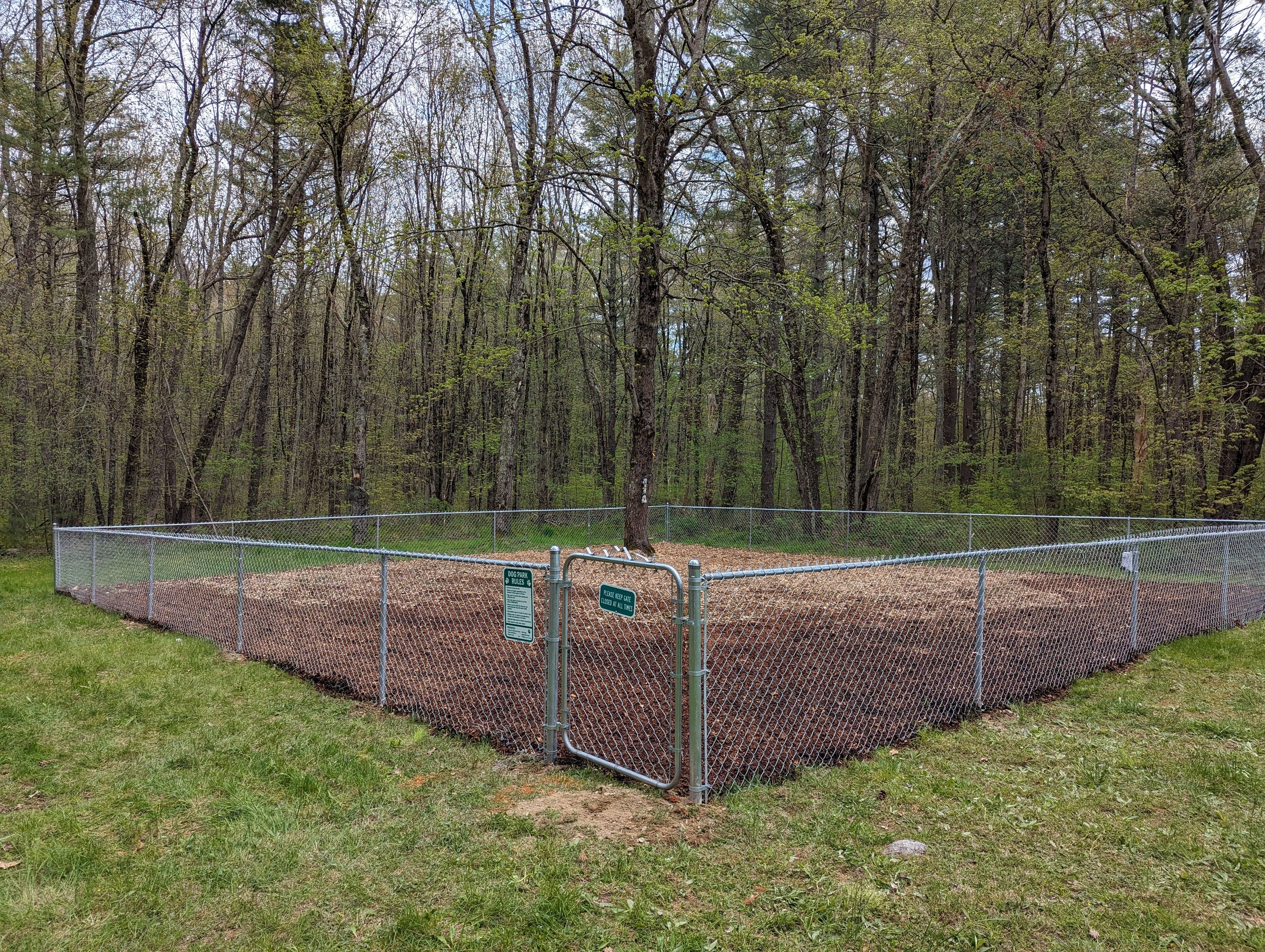A fenced-off area with freshly tilled soil, situated in a grassy area next to a forest with trees and budding leaves.