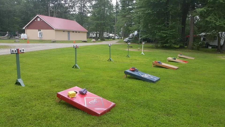 Outdoor cornhole game setup on a grassy area with several boards and score stands, surrounded by trees and a building in the background.