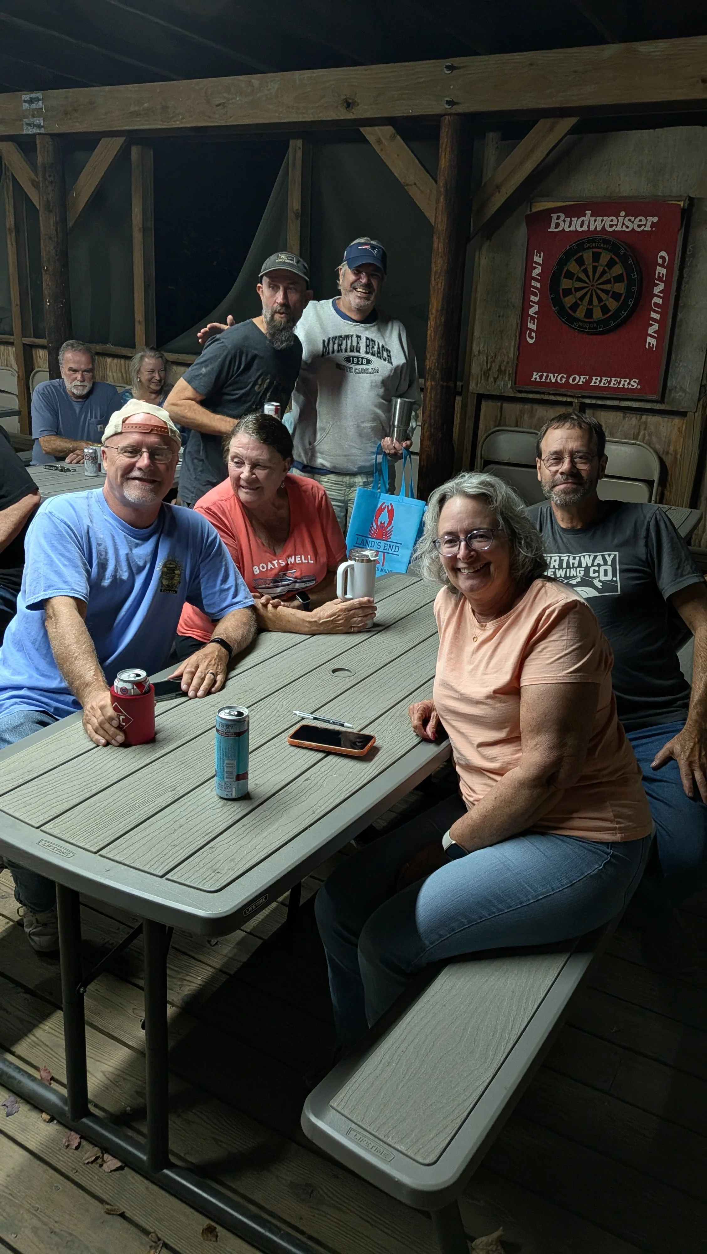 A group of six smiling adults gathered around a picnic table indoors, some holding drinks and a blue Land's End bag, with a Budweiser dartboard in the background.
