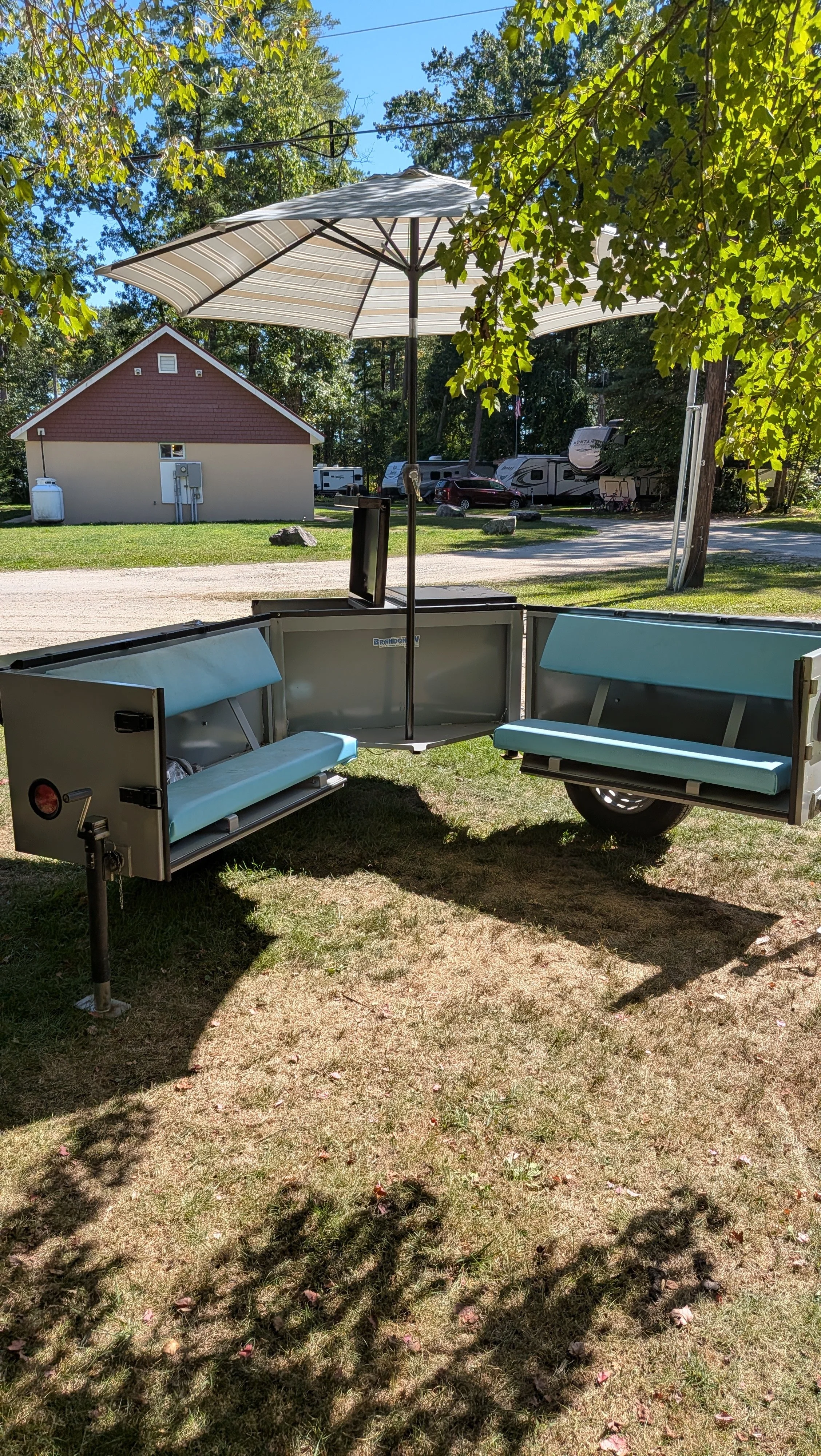 Outdoor scene with a vintage camper trailer featuring light blue cushions, a large striped umbrella, and a grassy area with trees and a building in the background.