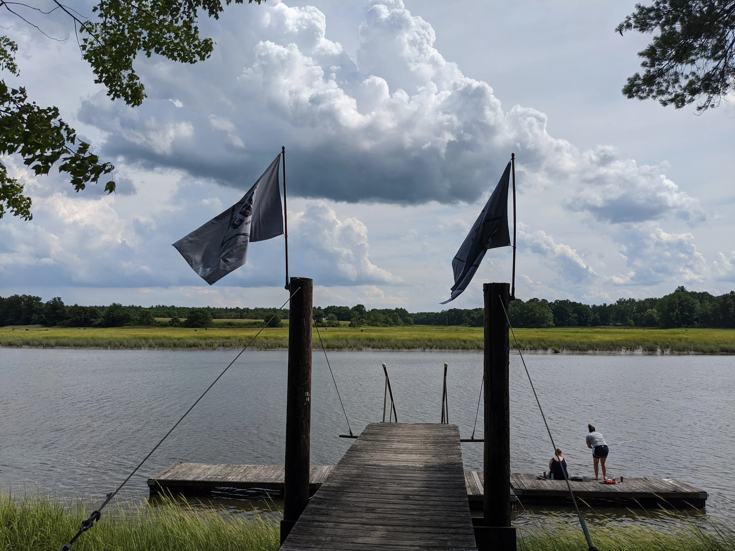 A wooden dock with two people sitting at the end, overlooking a river. Two flags are mounted on poles on the dock, and lush green trees and grass are visible along the riverbank, with a partly cloudy sky overhead.