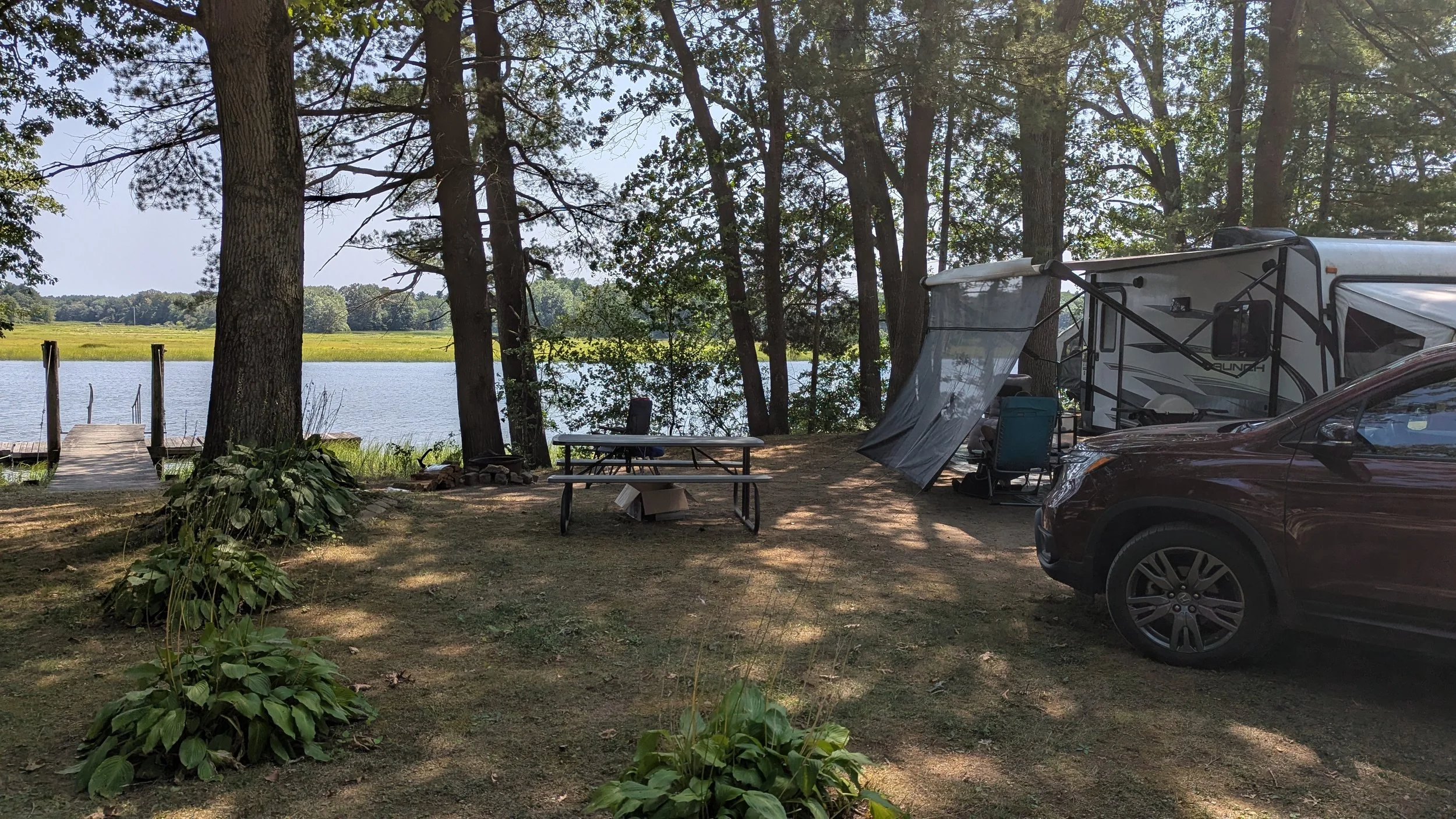 A lakeside camping scene with a camper trailer, a car, a picnic table, and a dock leading to the water, surrounded by trees.