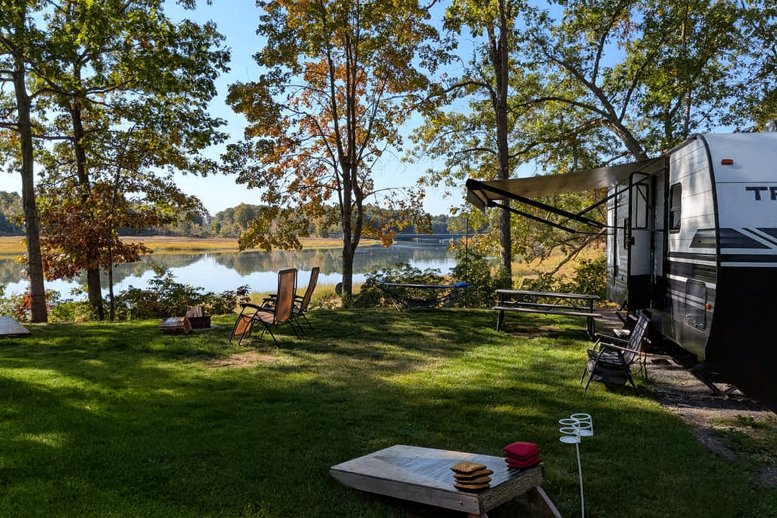 A lakeside campsite with a recreational vehicle, folding chairs, and a hammock surrounded by trees and lush grass, overlooking a calm lake.
