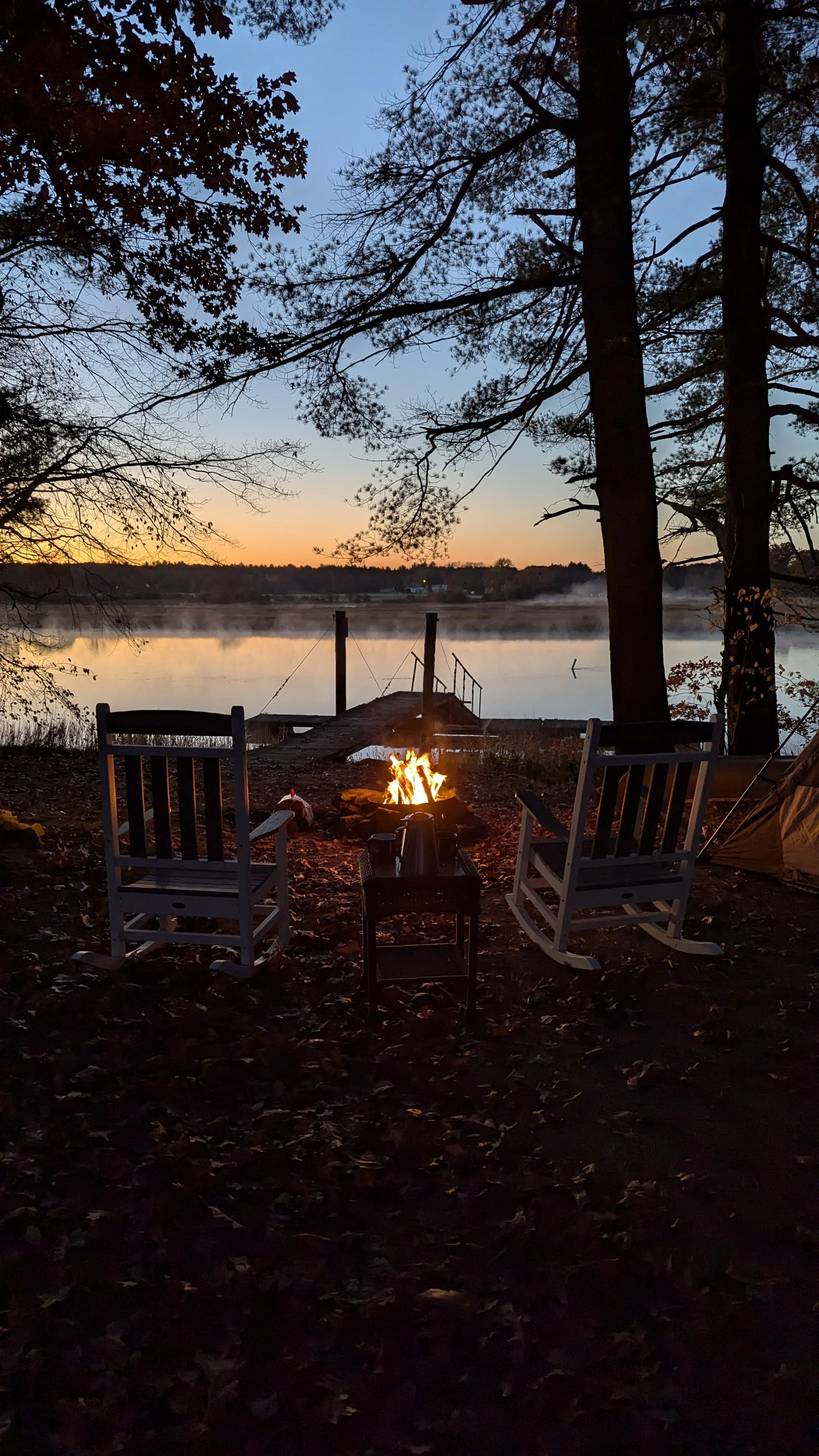 A peaceful lakeside scene at dusk with two white rocking chairs facing a campfire, a small table with a thermos and mugs, a tent to the right, a wooden dock extending over the water, and trees surrounding the area, with sunset colors in the sky and m