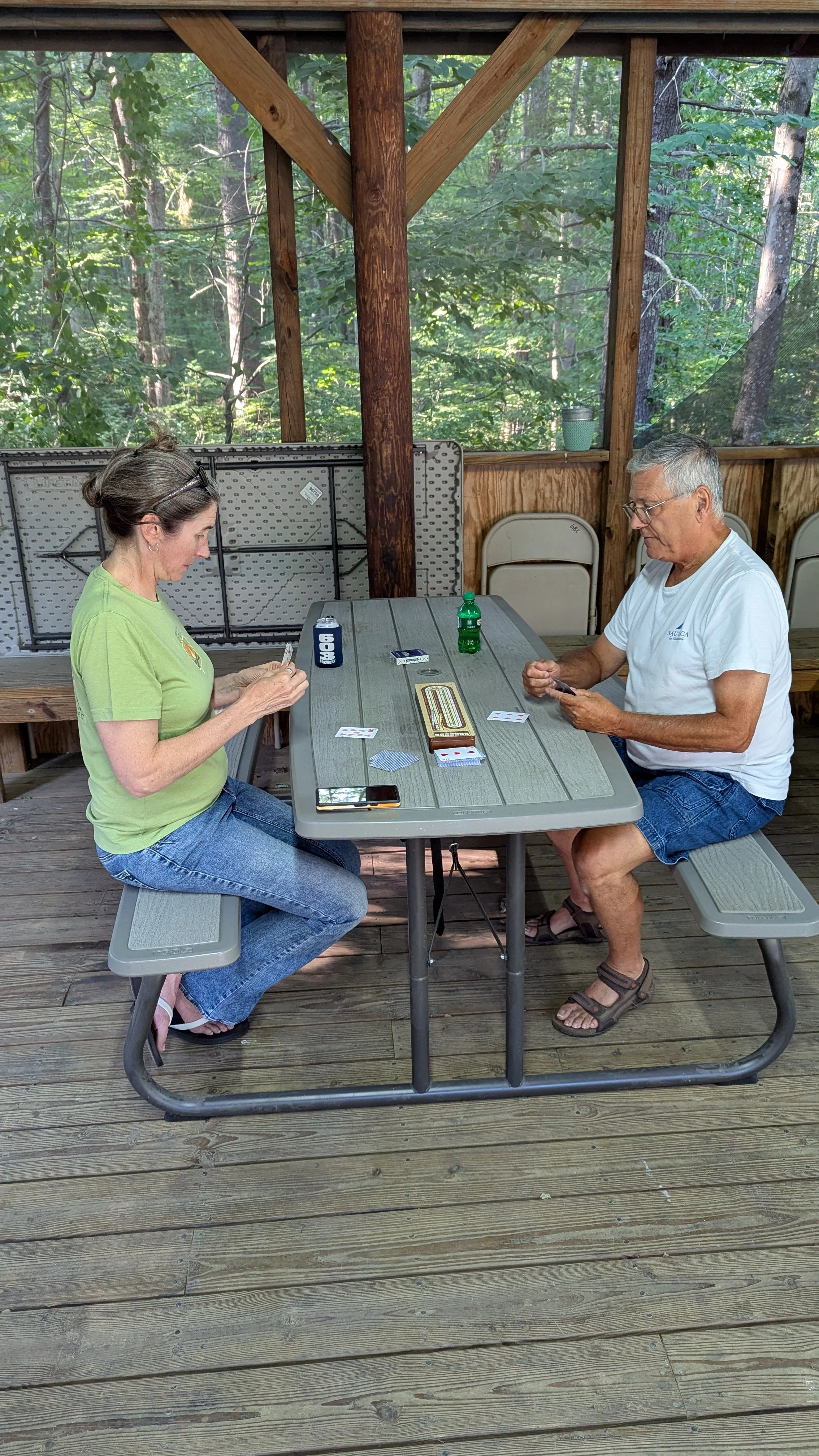 A man and woman sitting at a picnic table playing a card game in a wooded outdoor shelter.