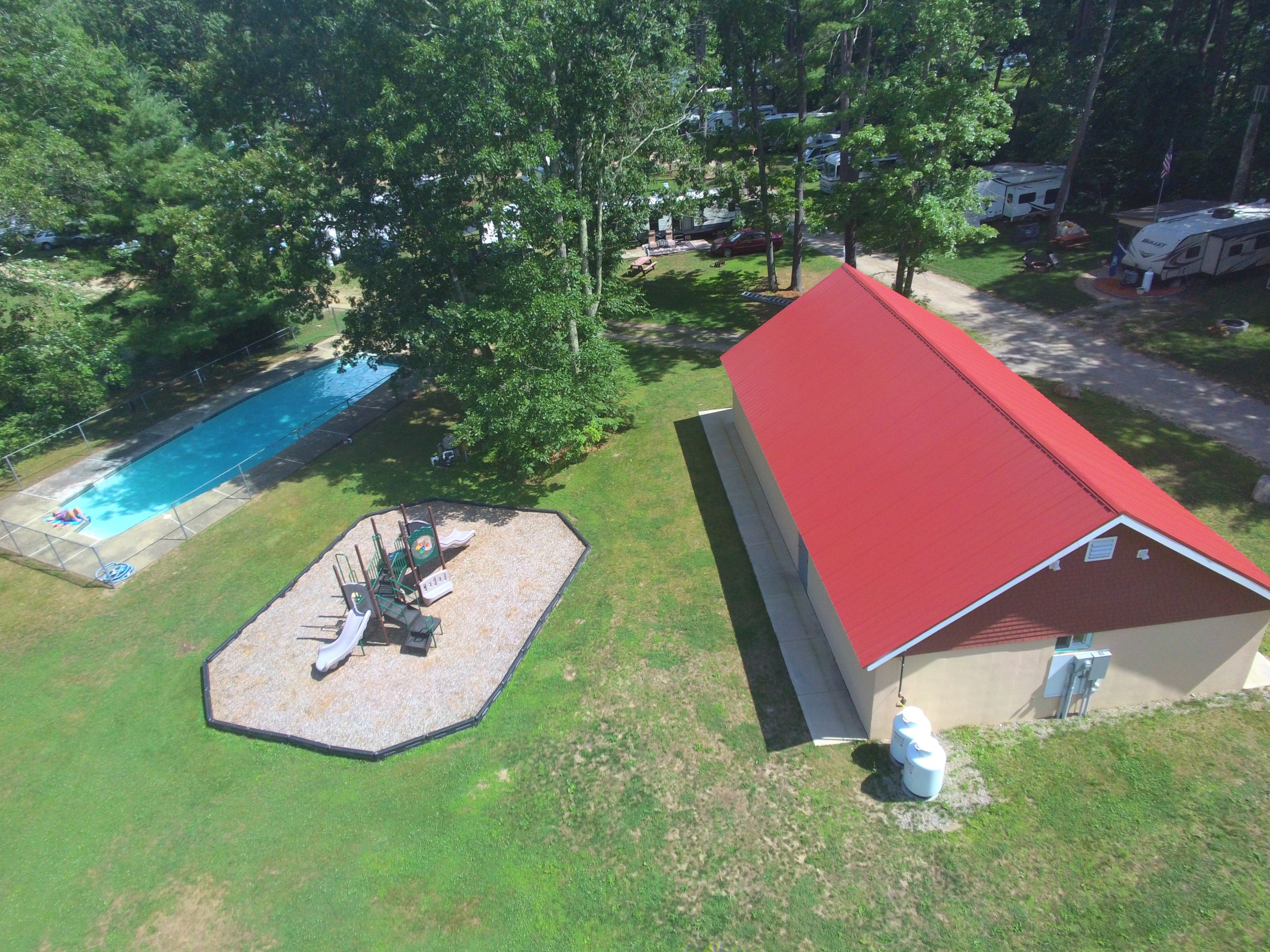 An aerial view of a backyard with a swimming pool, a playground, a red-roofed building, tall trees, and a parking area with RVs and cars.