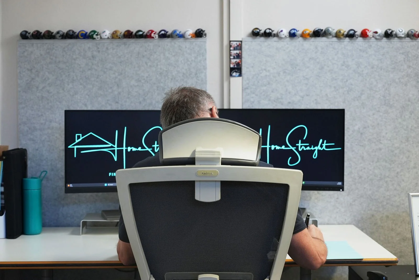 A man working at a desk in an office space, viewed from behind, with dual monitors displaying a logo that reads 'HomeStraight' in stylized text.