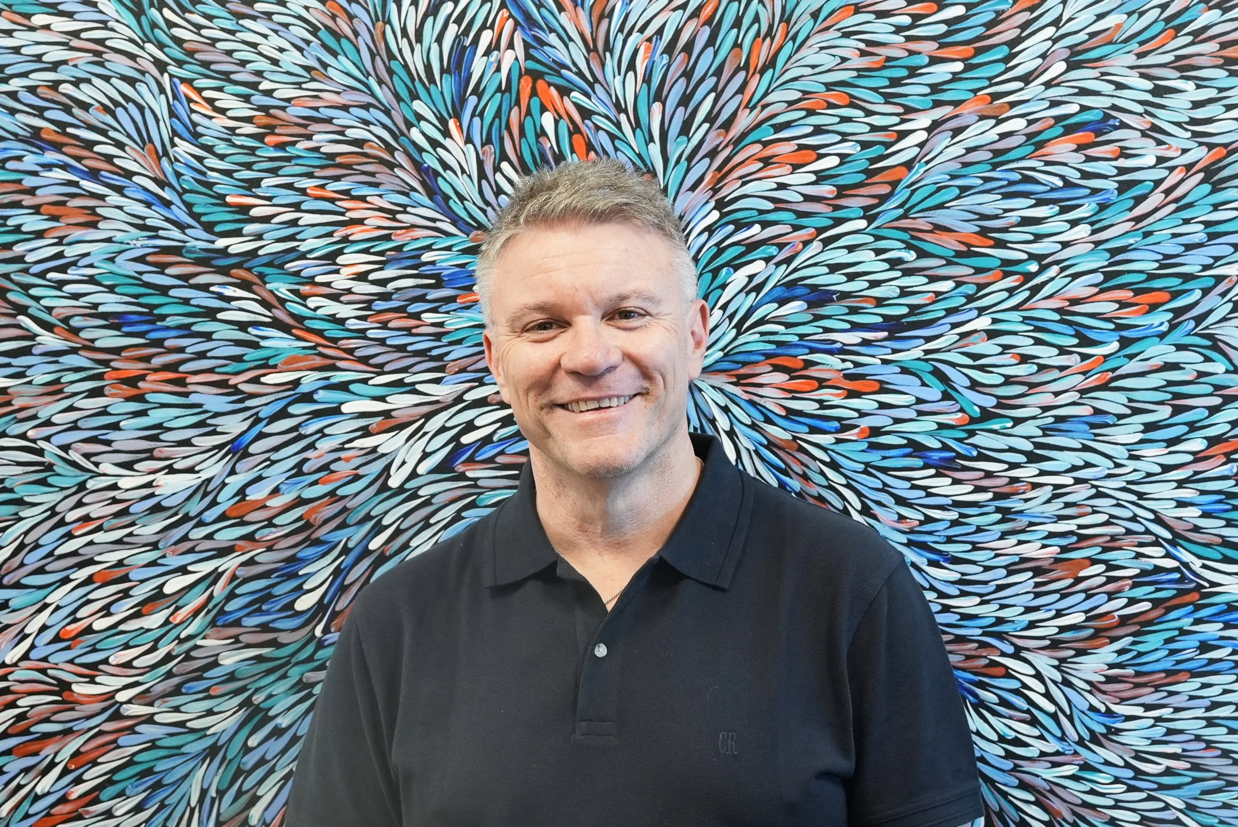 A man smiling, standing in front of an abstract colorful mural with swirl patterns of red, blue, black, white, and gray.
