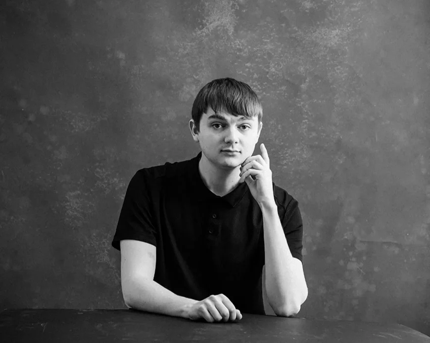 A young man with short hair and a thoughtful expression, wearing a black shirt, sitting at a table against a textured wall.