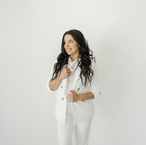A woman with long dark wavy hair wearing a white blazer and matching pants standing against a plain white background.