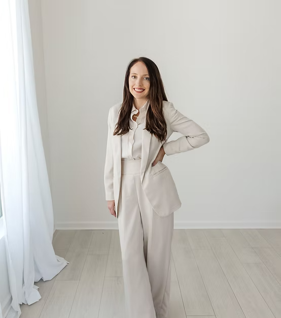 A woman wearing a white pantsuit standing in a bright, minimalistic room with white walls and light-colored flooring, smiling at the camera.
