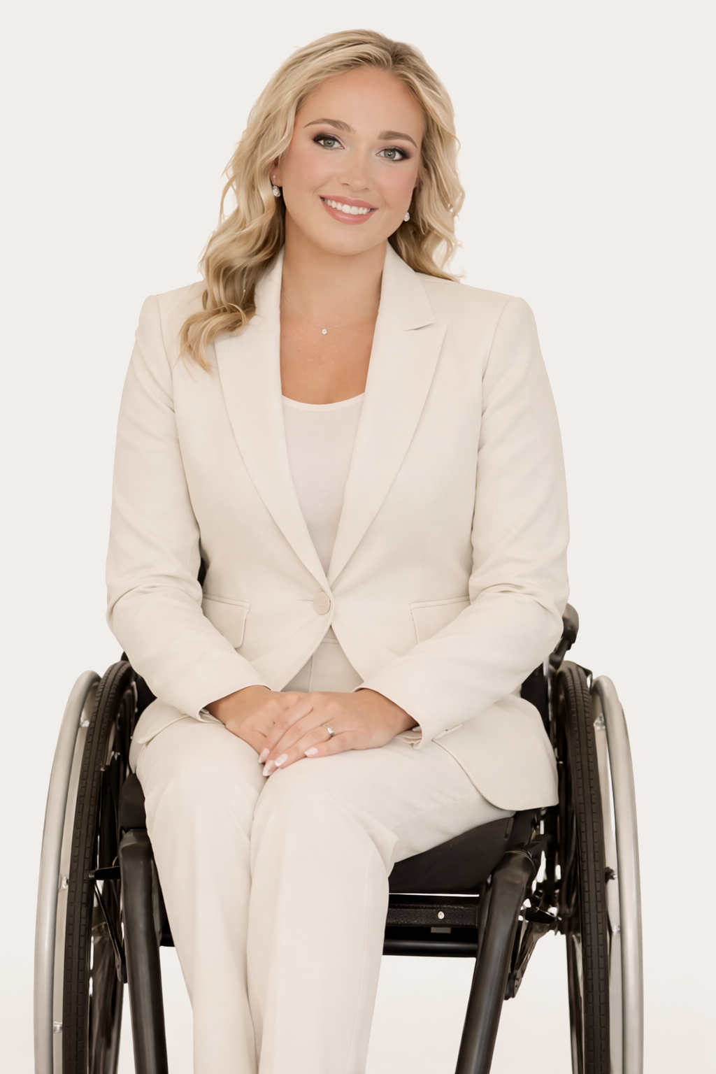 A woman in a white suit sitting in a wheelchair, smiling with blonde curly hair against a plain white background.