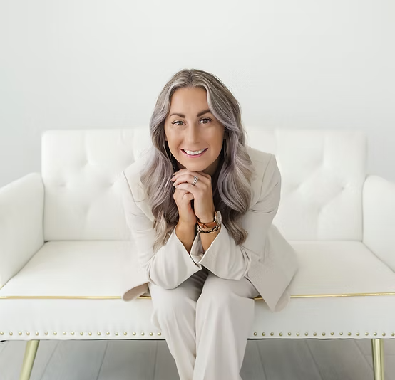 A smiling woman with long gray hair sitting on a white couch in a bright, minimalistic room.