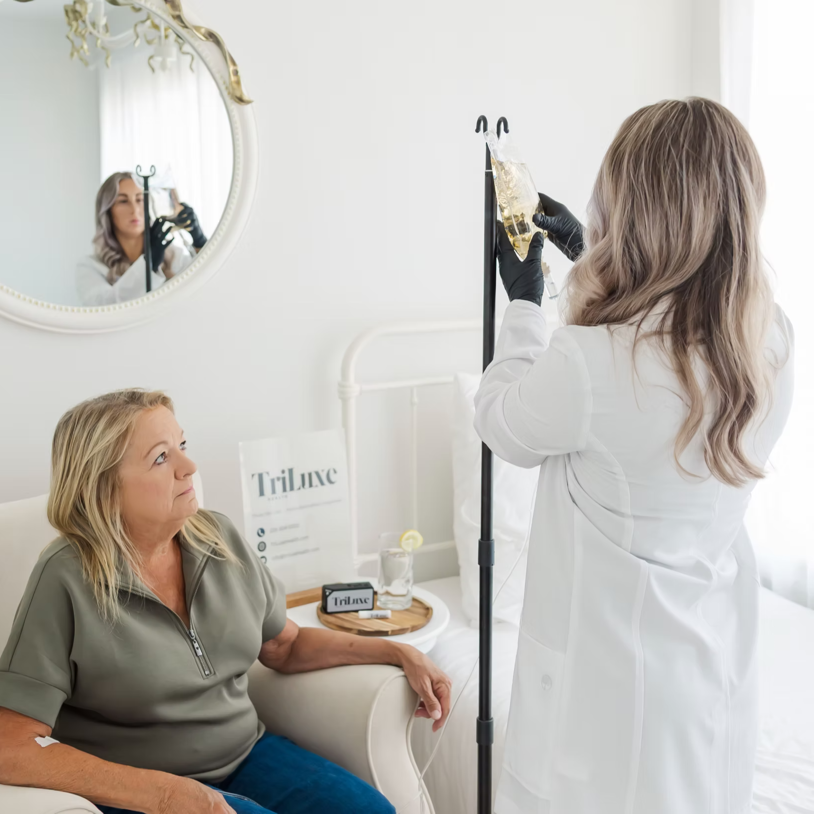 A woman with blonde hair sitting in a hospital bed, receiving IV fluids from a healthcare professional. The healthcare worker, dressed in white, is preparing the IV drip, which contains a clear liquid. The patient is looking at the healthcare worker, and her hand is resting on a tray with a branded card and a glass of water. A mirror on the wall reflects the scene.
