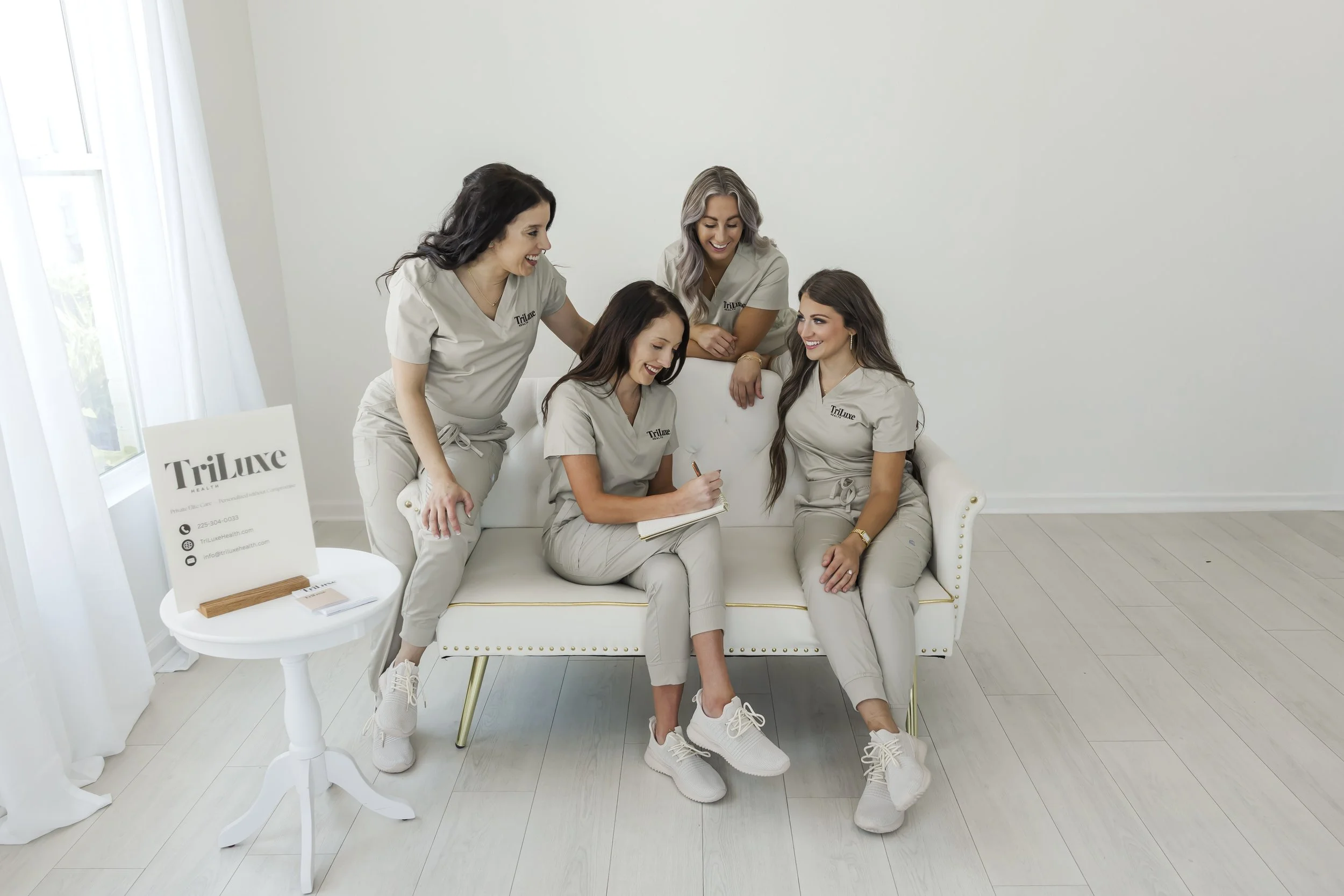 Five women in matching beige scrubs sitting and standing inside a bright, minimalistic room with white walls and light-colored flooring. They are smiling, chatting, and one is writing in a notebook. A small round table holds a sign with business information and a few products. The women appear to be part of a professional team, possibly in healthcare or beauty services.