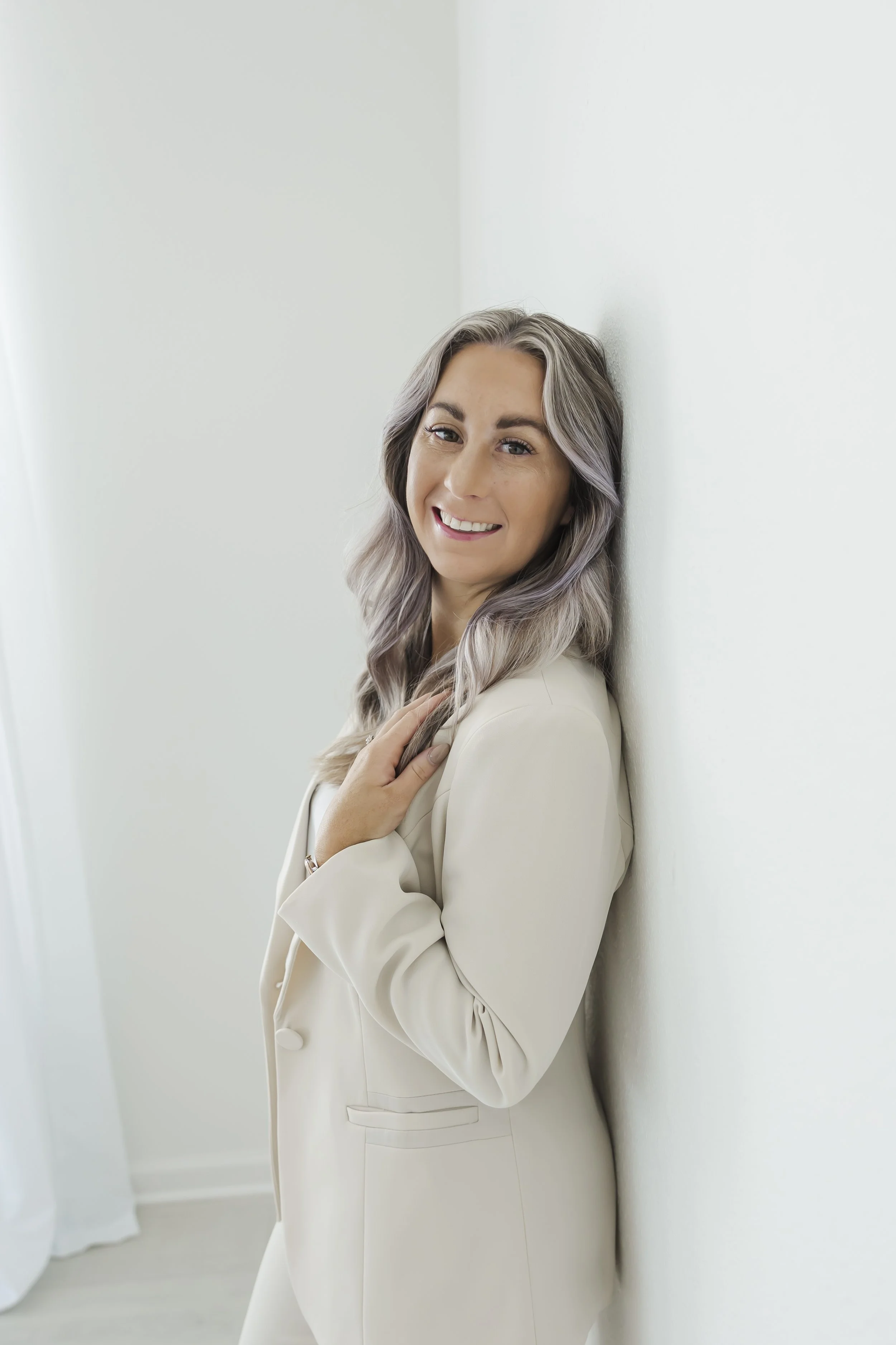 A woman with gray, wavy hair smiling in a white blazer, standing against a white wall.
