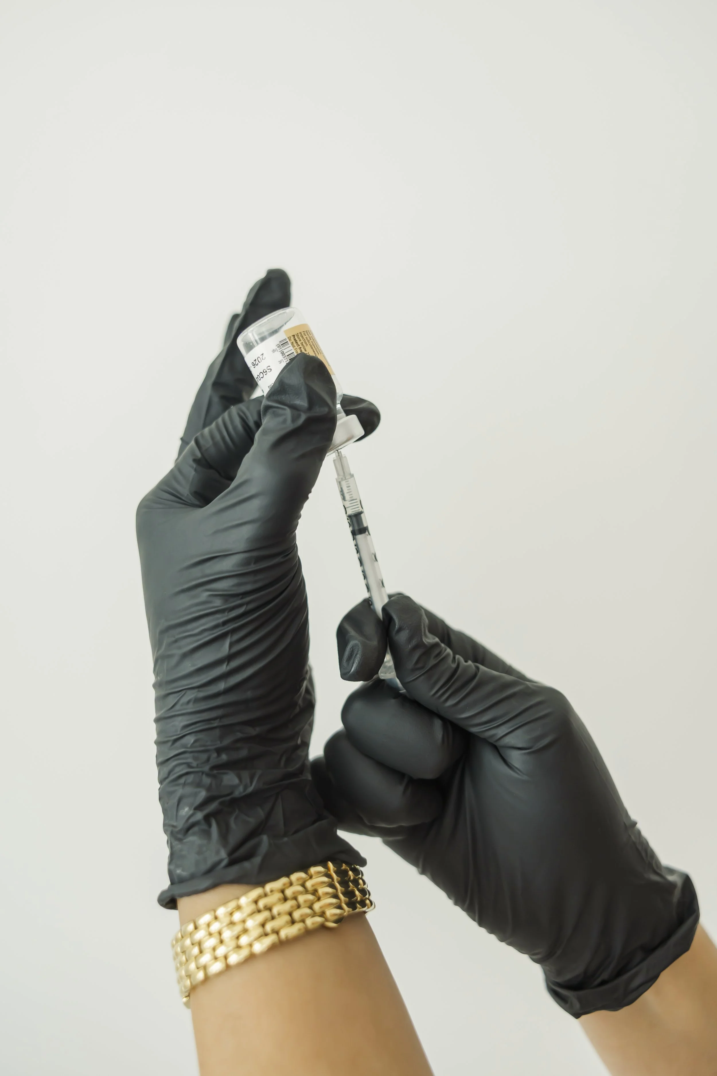 Person wearing black gloves holding a syringe and a small vial, with a gold bracelet on wrist, against a plain white background.