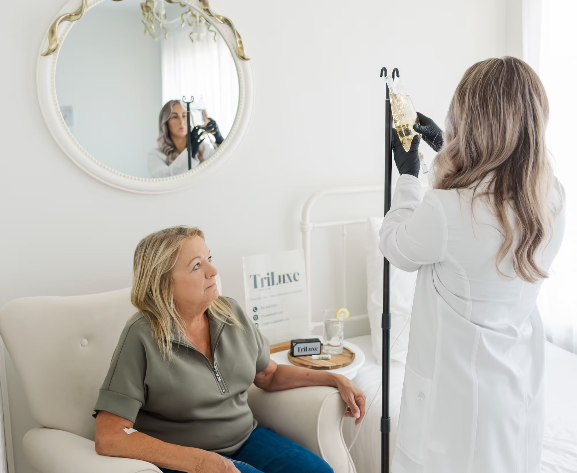 A woman in a grey shirt sits on a white chair in a bright room with white walls and looks up at a healthcare professional preparing to draw her blood, with a mirror reflecting the woman taking a photo.