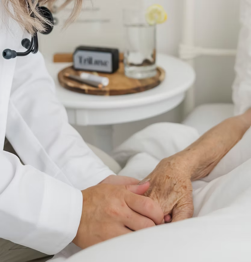 A healthcare professional holding an elderly patient's hand in a gentle, comforting manner in a bright bedroom.