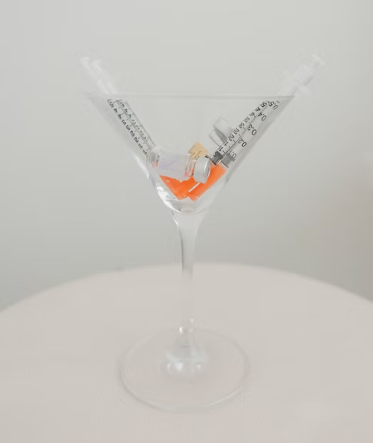 A cocktail glass with a clear liquid, holding two syringes and a small orange pill.