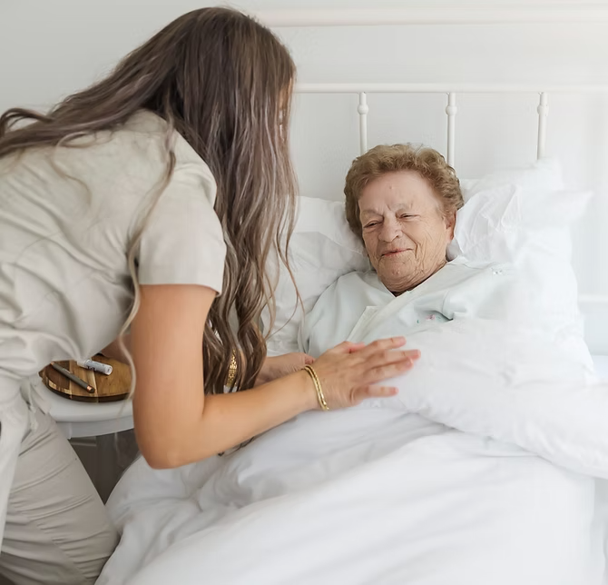 A young woman with long brown hair leaning over a bed to comfort an elderly woman in a hospital bed, with white bedding and a white headboard.