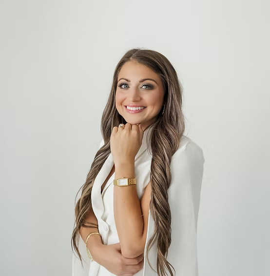 A young woman with long wavy brown hair, wearing a white blazer, smiling, with a gold bracelet and watch, posing against a plain light gray background.