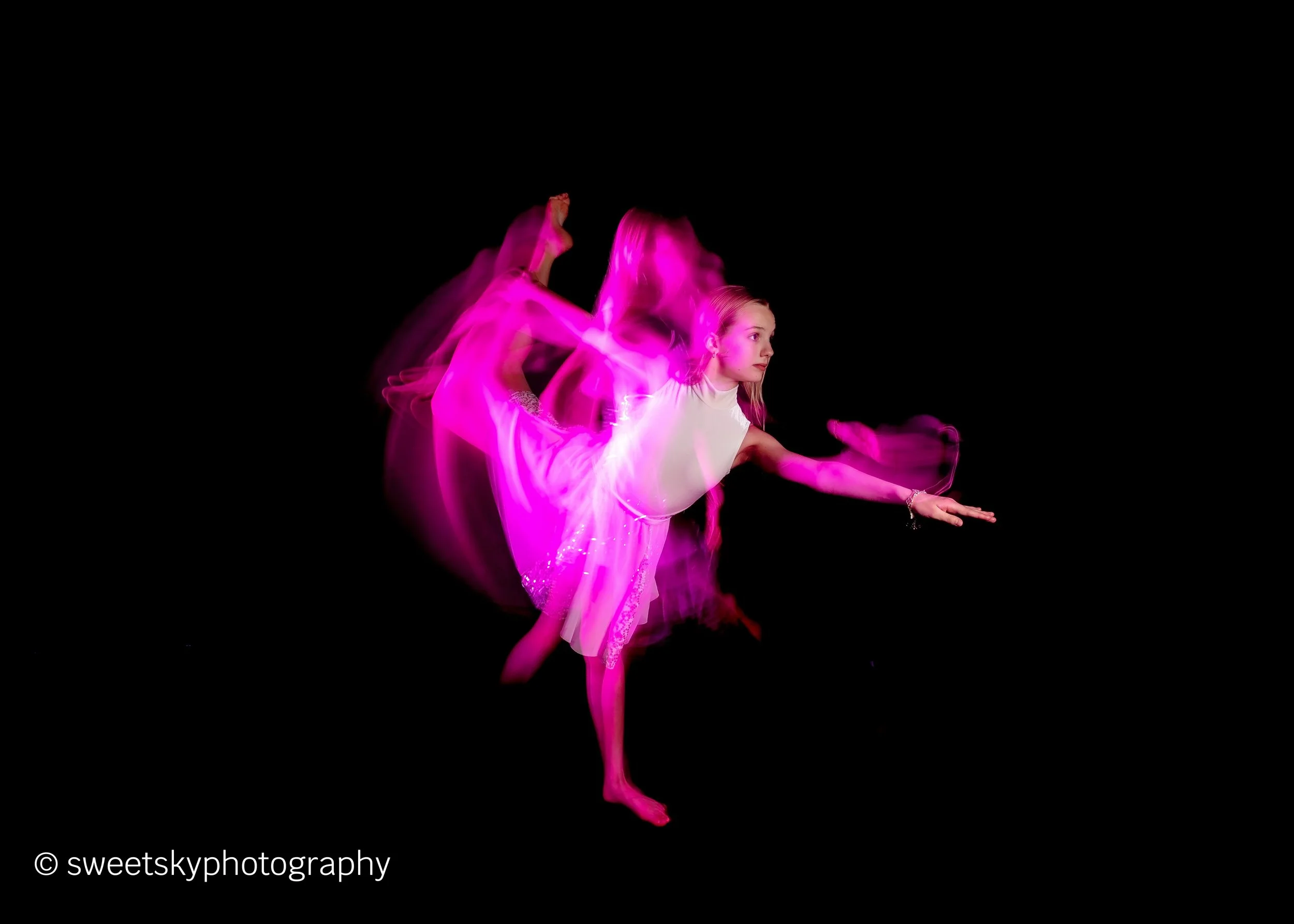Young girl in a white dress performing a dance move against a black background, with pink motion blur effects.