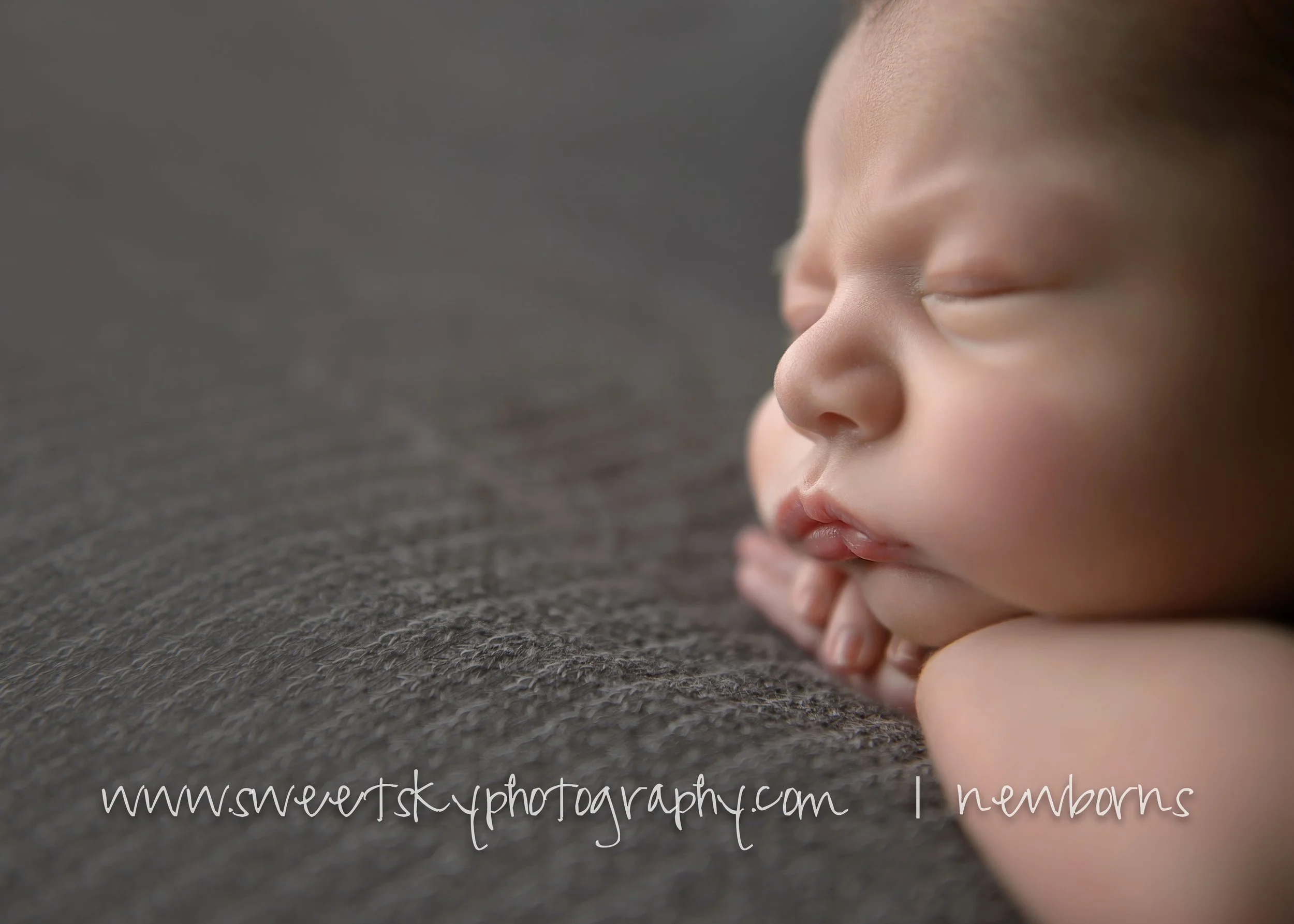 Close-up of a sleeping newborn baby's face resting on a dark textured surface with text overlay 'www.sweertskyphotography.com | 1 newborns'