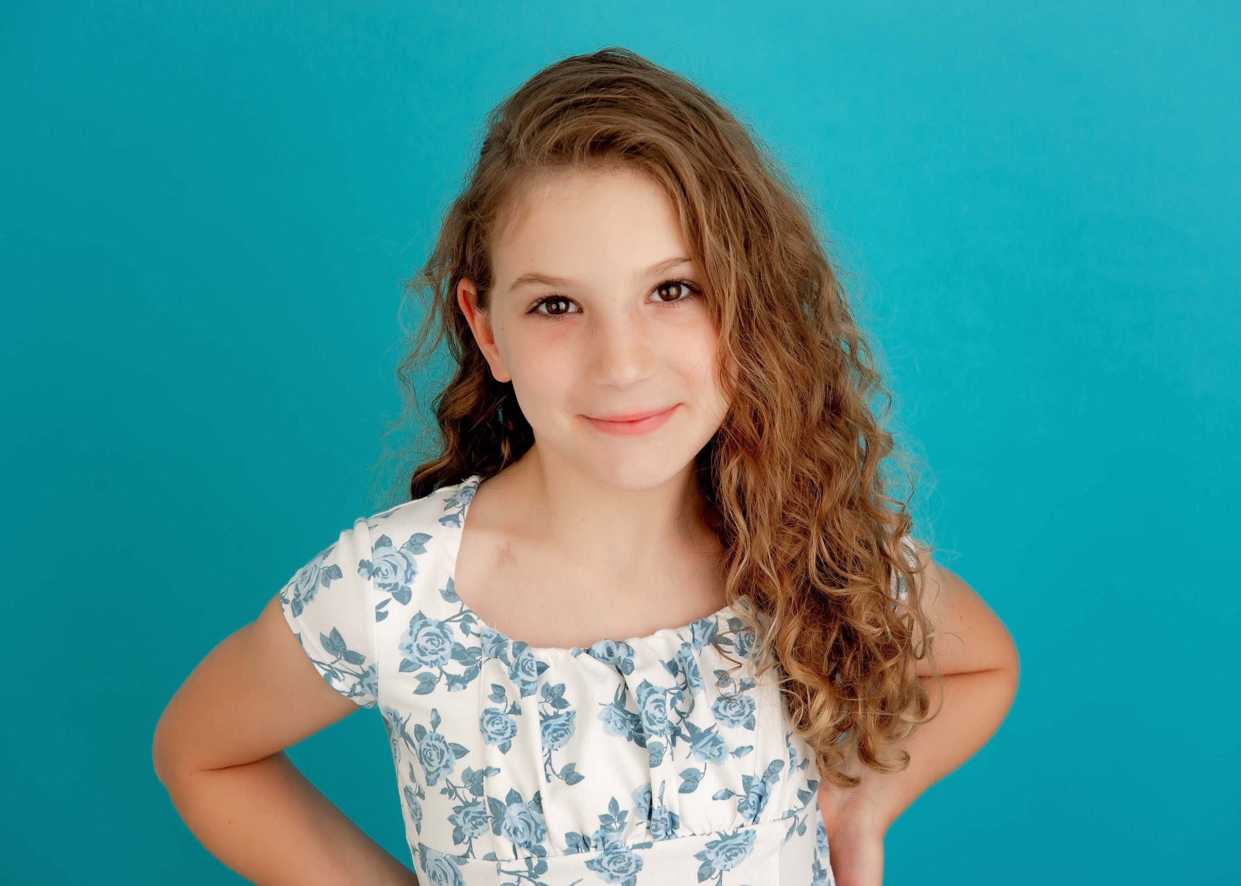 A young girl with long, curly brown hair and a gentle smile stands against a bright turquoise background. She is wearing a white dress with a blue floral pattern and has her hands on her hips.