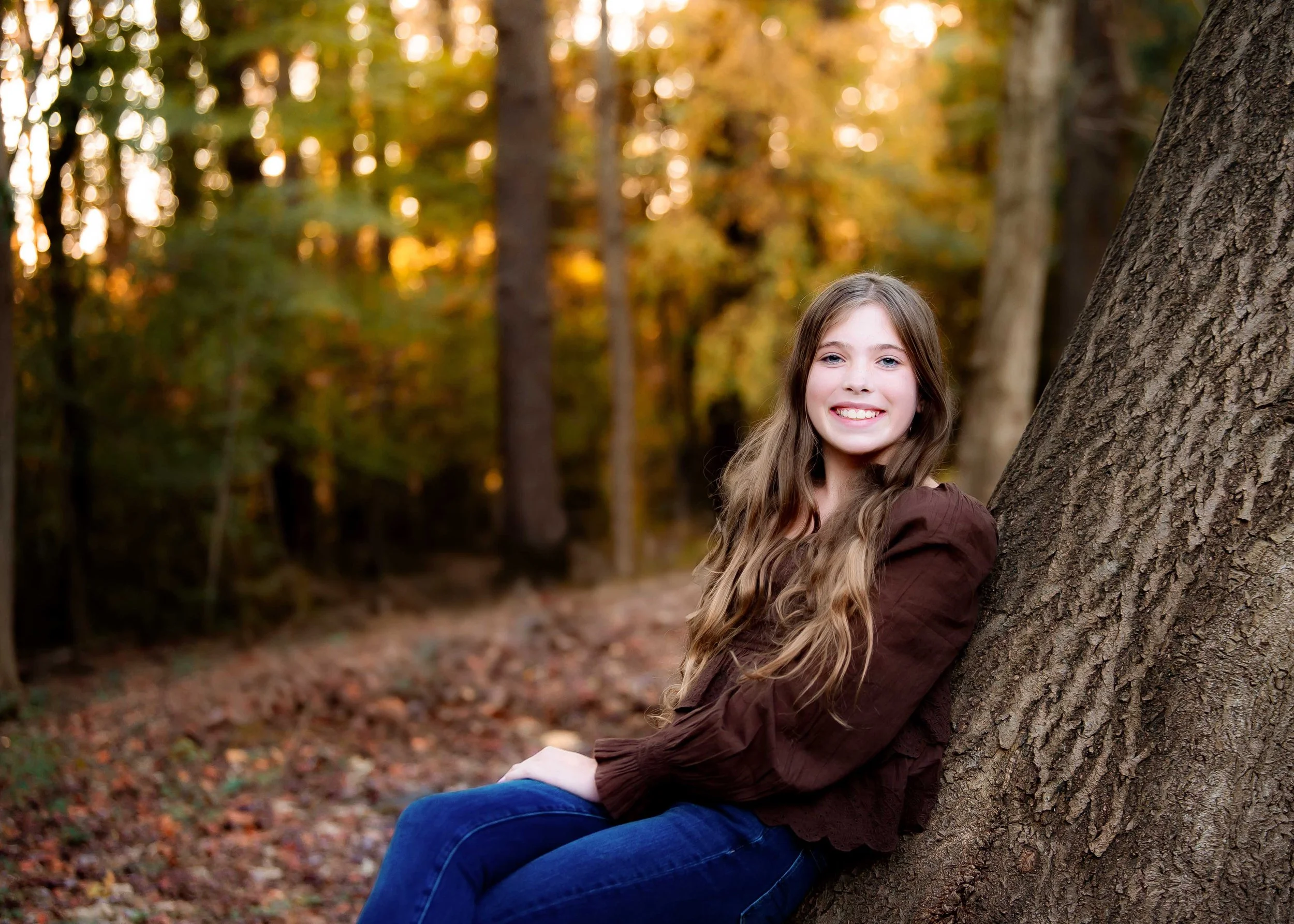 A young girl with long wavy hair sitting on the ground and leaning against a tree in the woods during autumn, smiling at the camera with fall foliage in the background.