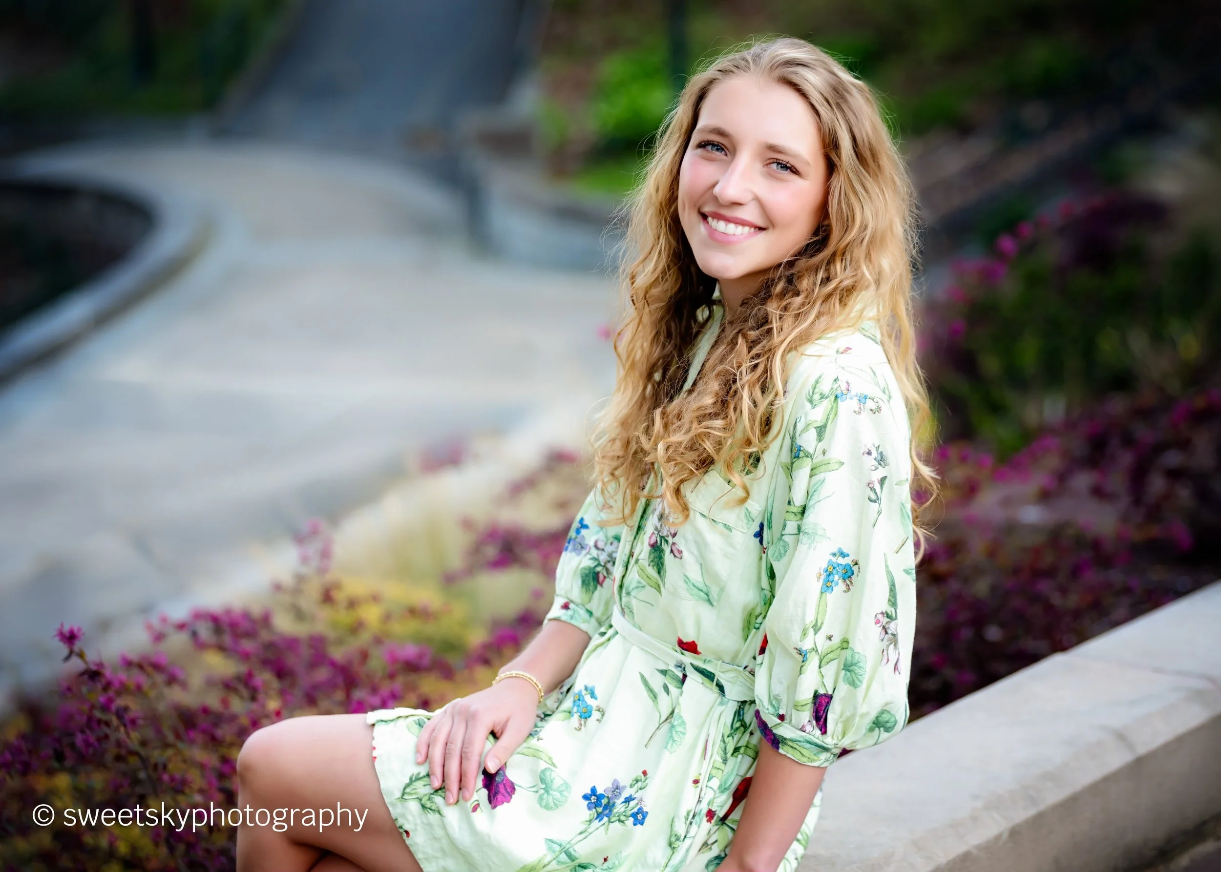 A young woman with long, wavy blonde hair, smiling and sitting outdoors on a concrete ledge, wearing a light yellow dress with a floral pattern, in a park with greenery and pink flowers.