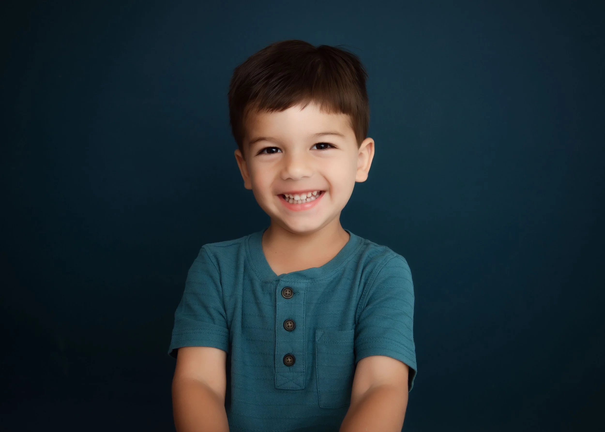 A young boy with dark hair and a bright smile, wearing a teal shirt, standing against a dark background.