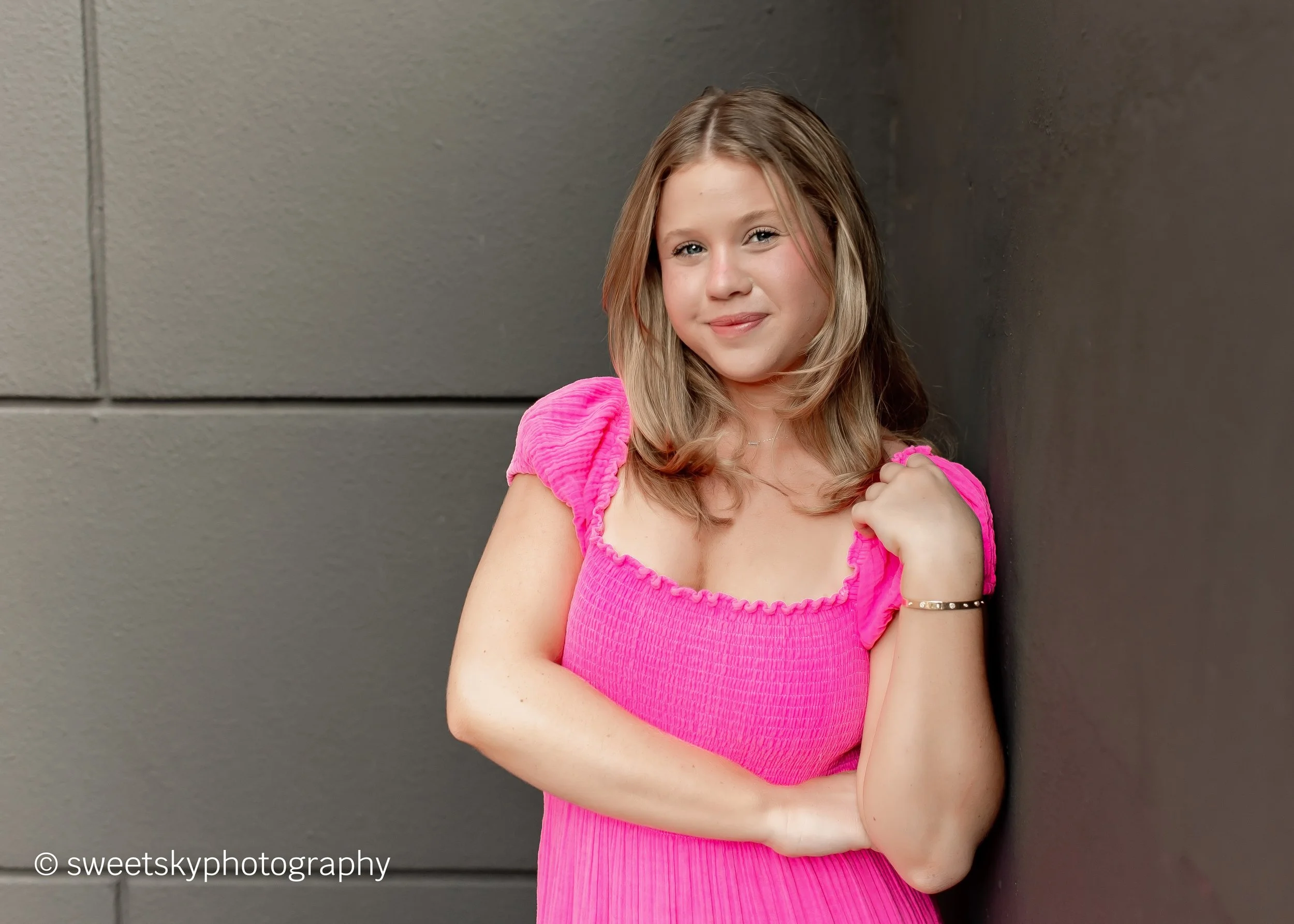A young girl with light brown hair and a pink dress posing against a gray wall, smiling at the camera.