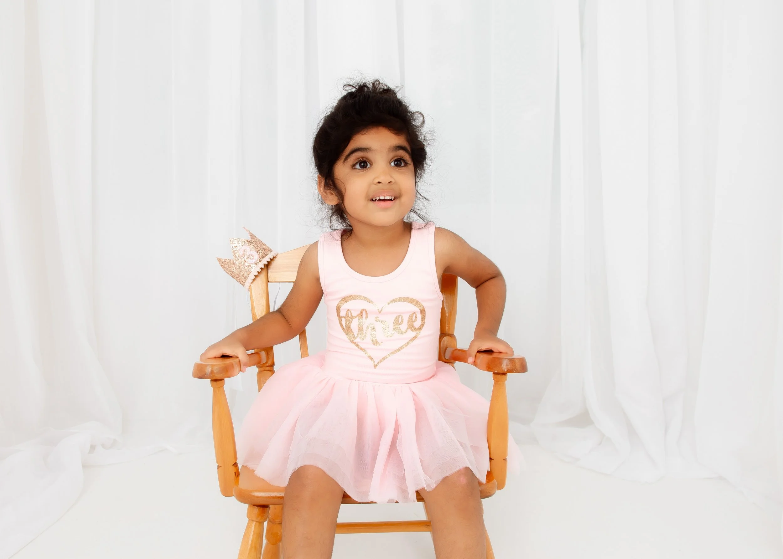 Young girl sitting on a wooden high chair, wearing a pink dress with 'free' written in gold, and a small pink crown on the back of her chair, in front of white curtains.