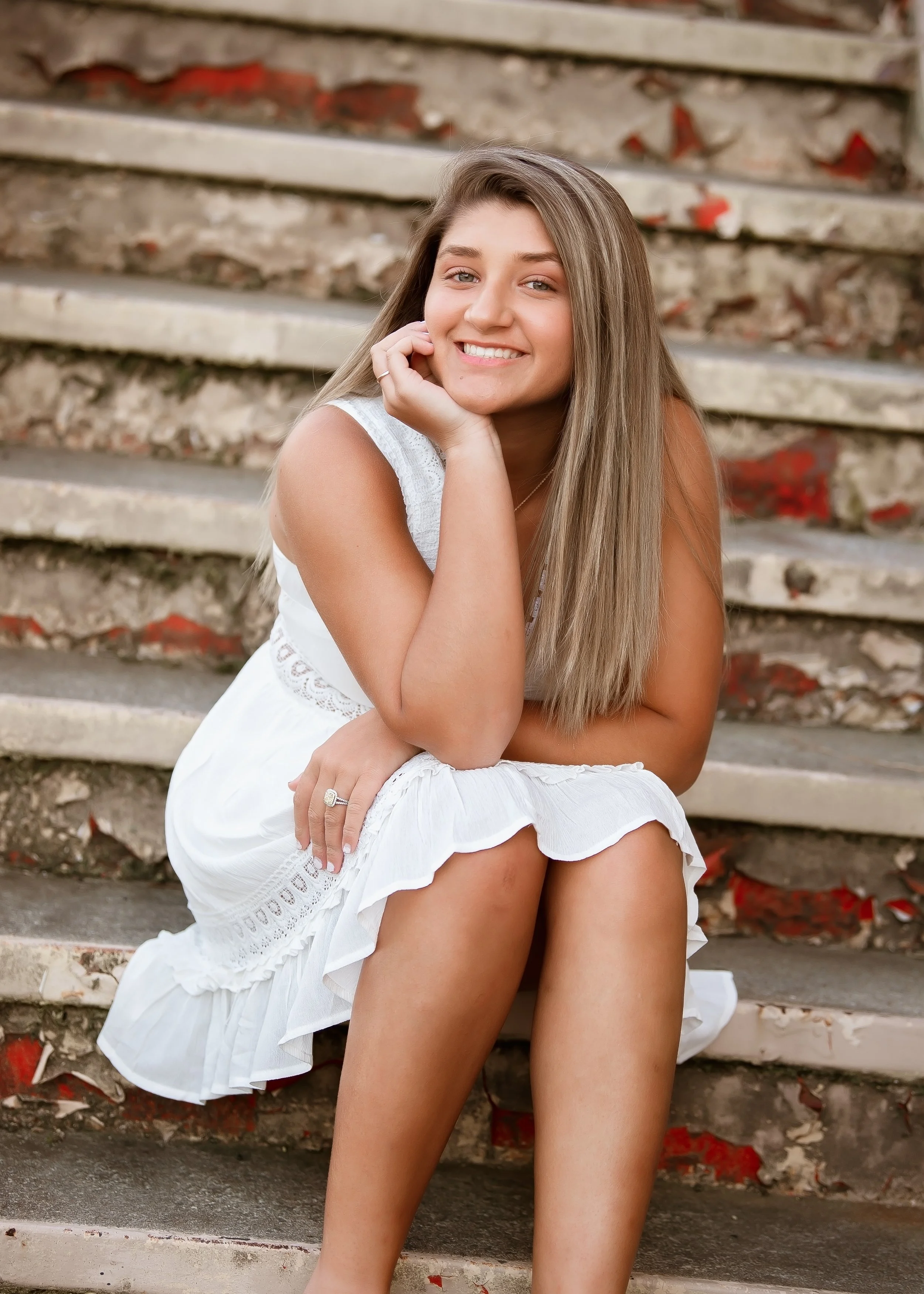 A young woman wearing a white dress, sitting on outdoor steps with a smile, hand resting on her chin, and long, straight hair.
