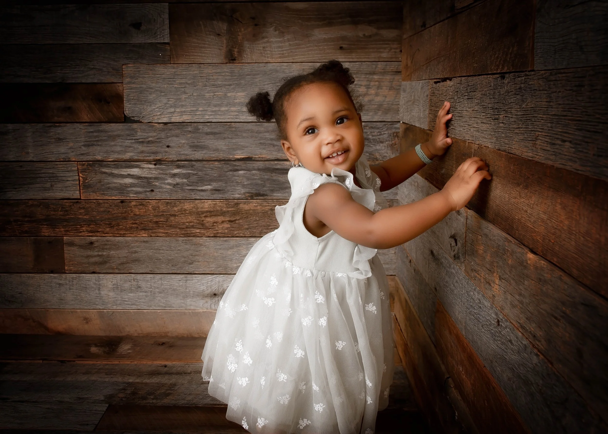 Smiling young girl in a white dress with ruffles, standing in a corner with wooden walls, looking up at the camera.