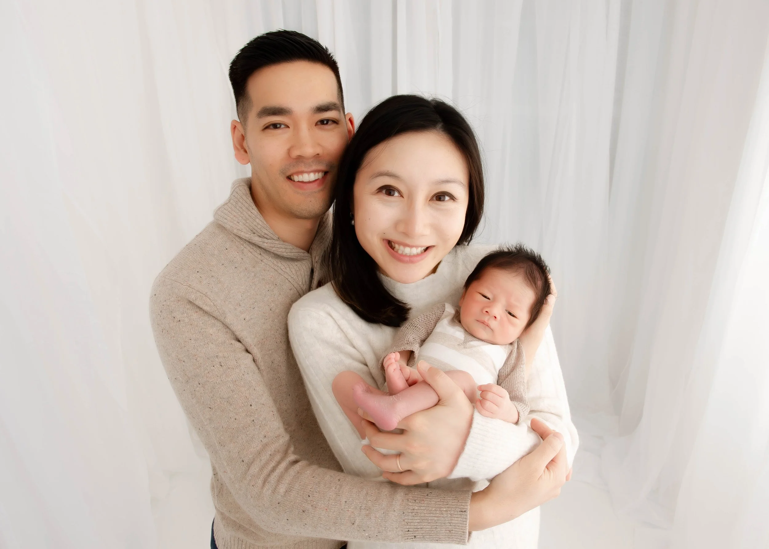 Family of three – a man, a woman, and a newborn baby – smiling and posing for a photo indoors with white curtains in the background.