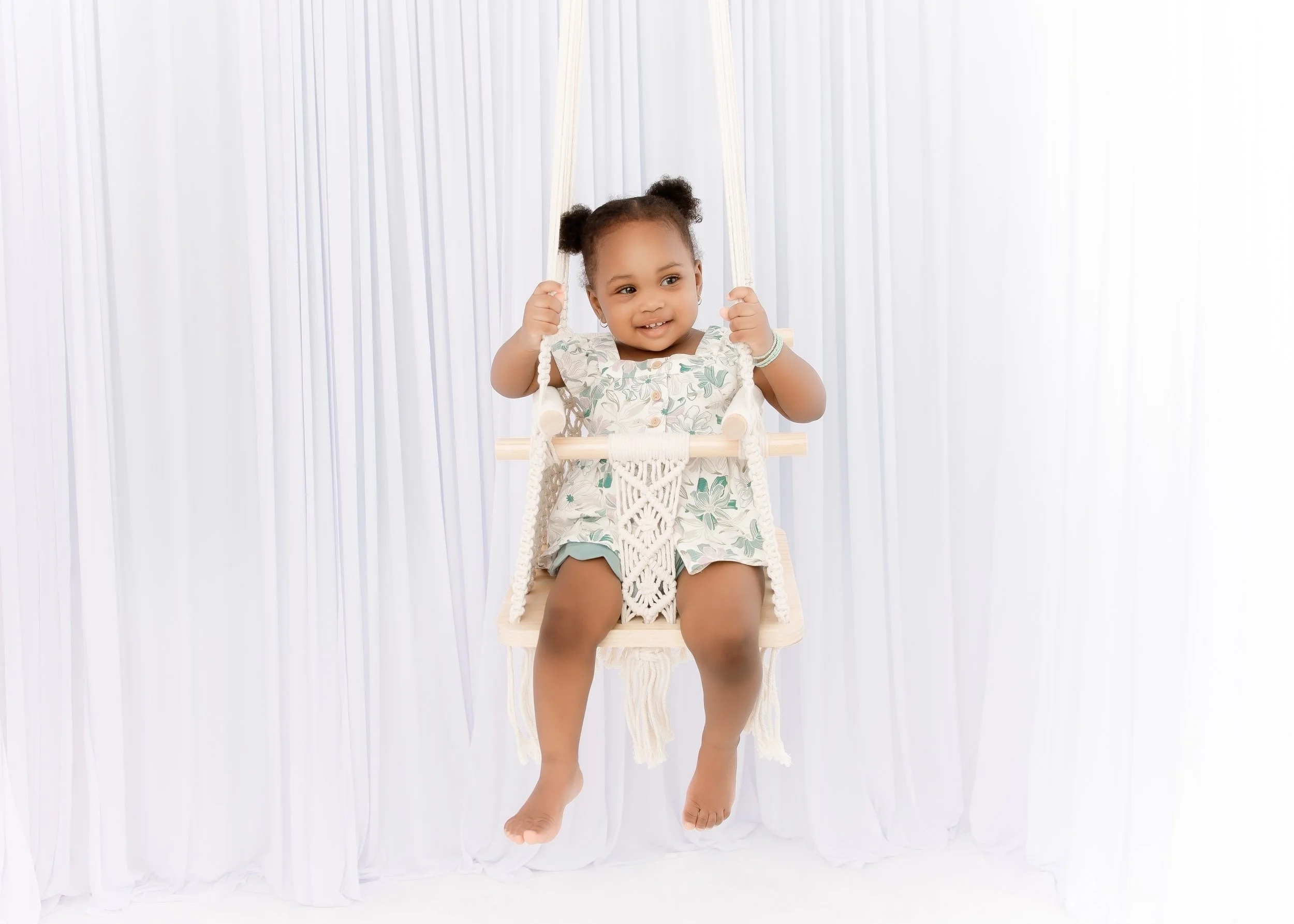 A young girl with pigtails sitting on a wooden swing with a white macrame cushion, smiling, in front of a white curtain background.
